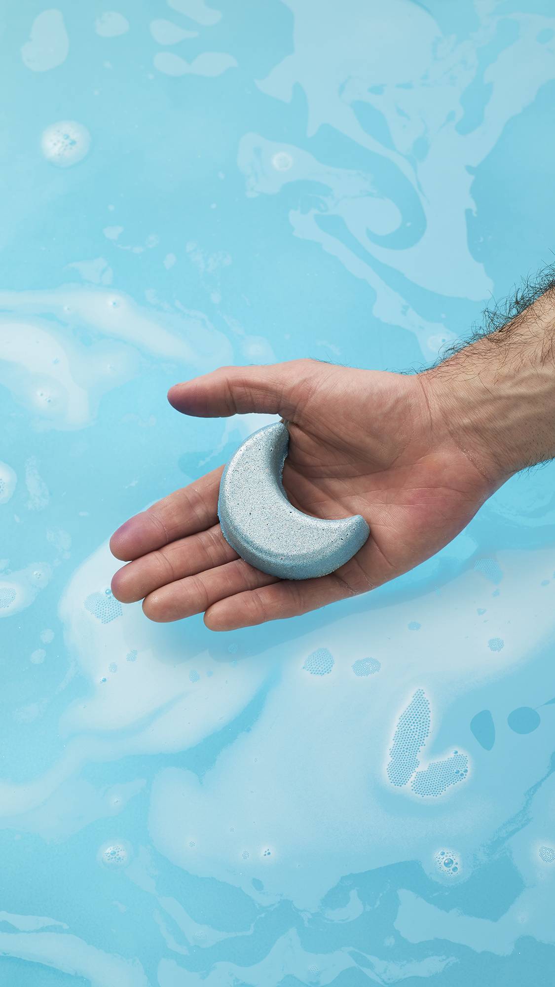 A person's open palm holds the shimmering, silver-blue Sukoon bath bomb, shaped like a crescent moon, over bright blue bath water topped with white foam and bubbles.