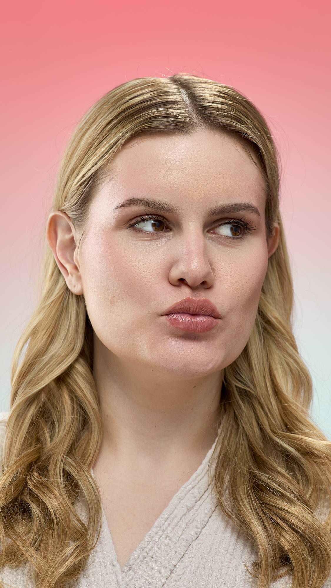 A person with blonde wavy hair pouts their lips, showcasing a glossy finish from Super Milk lip gloss. They are wearing a light, textured robe against a pink-to-blue gradient background.