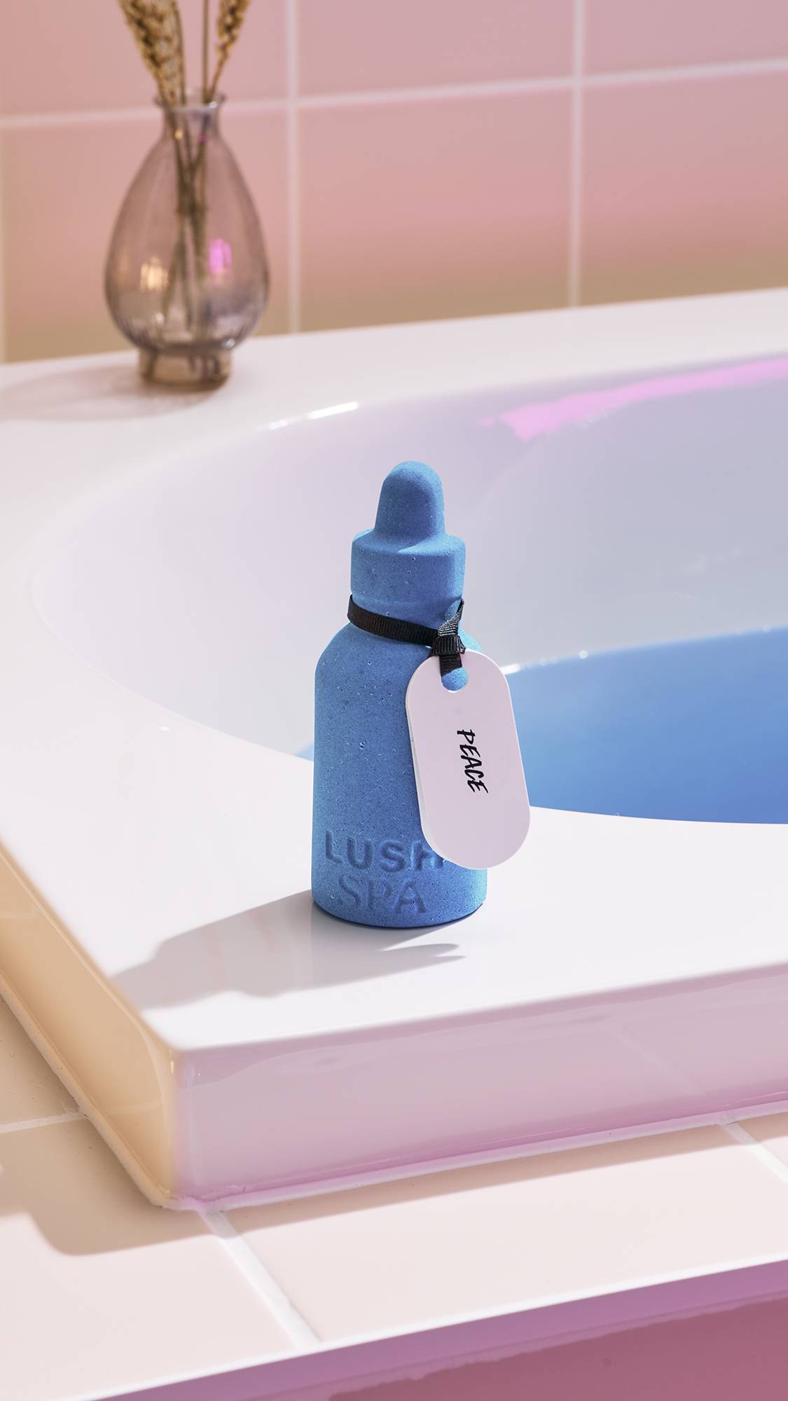 A vibrant blue bath bomb shaped like a dropper bottle sits on the edge of a white bathtub. It features an embossed 'LUSH SPA' logo and a white 'PEACE' tag tied around its neck, set against a background of soft pink tiles and a glass vase.