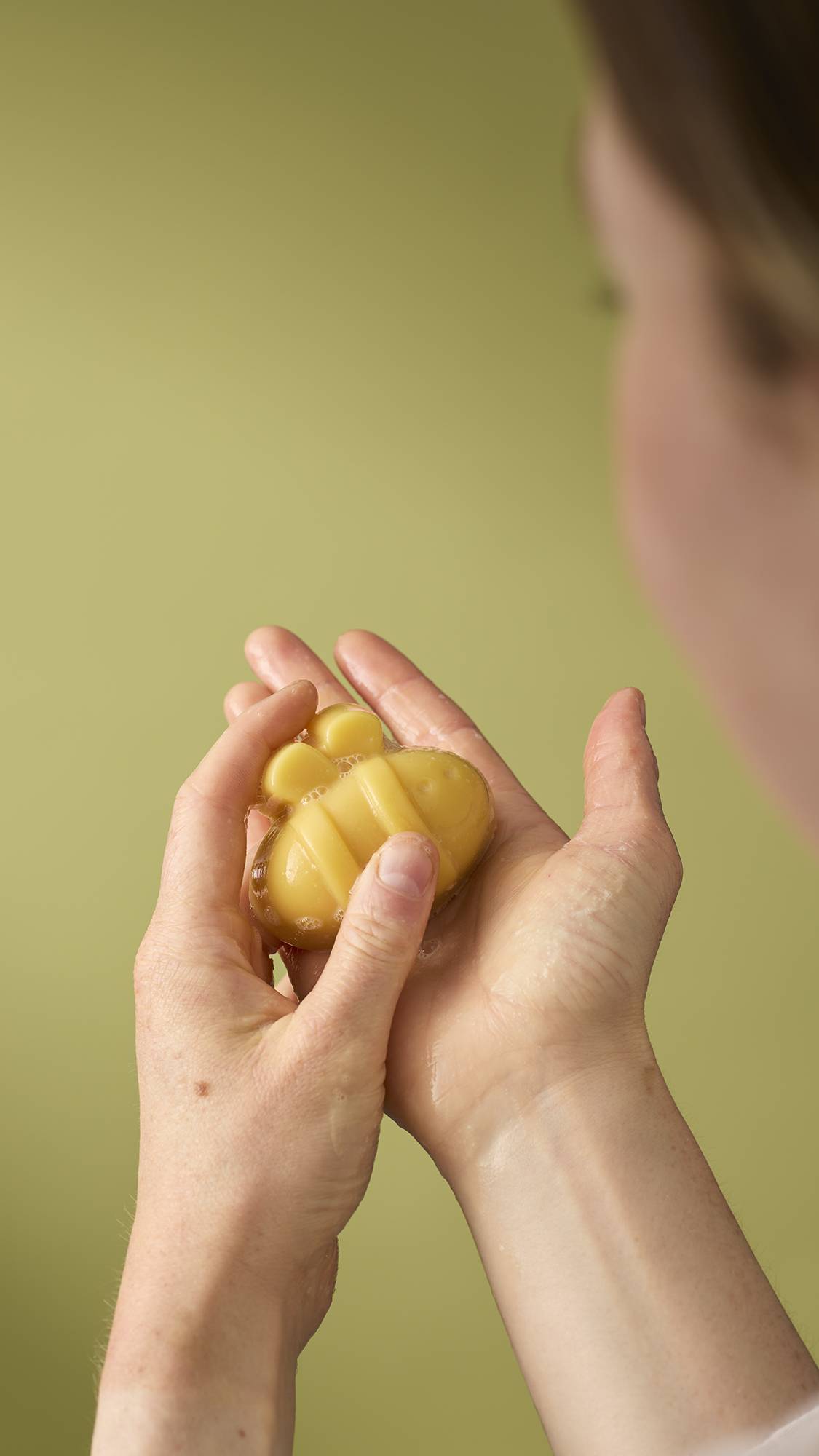 A person holds a small, yellow, bee-shaped facial gummy wash in their wet hands. The product is slightly bubbly, and the person appears to be lathering it against an olive-green background.