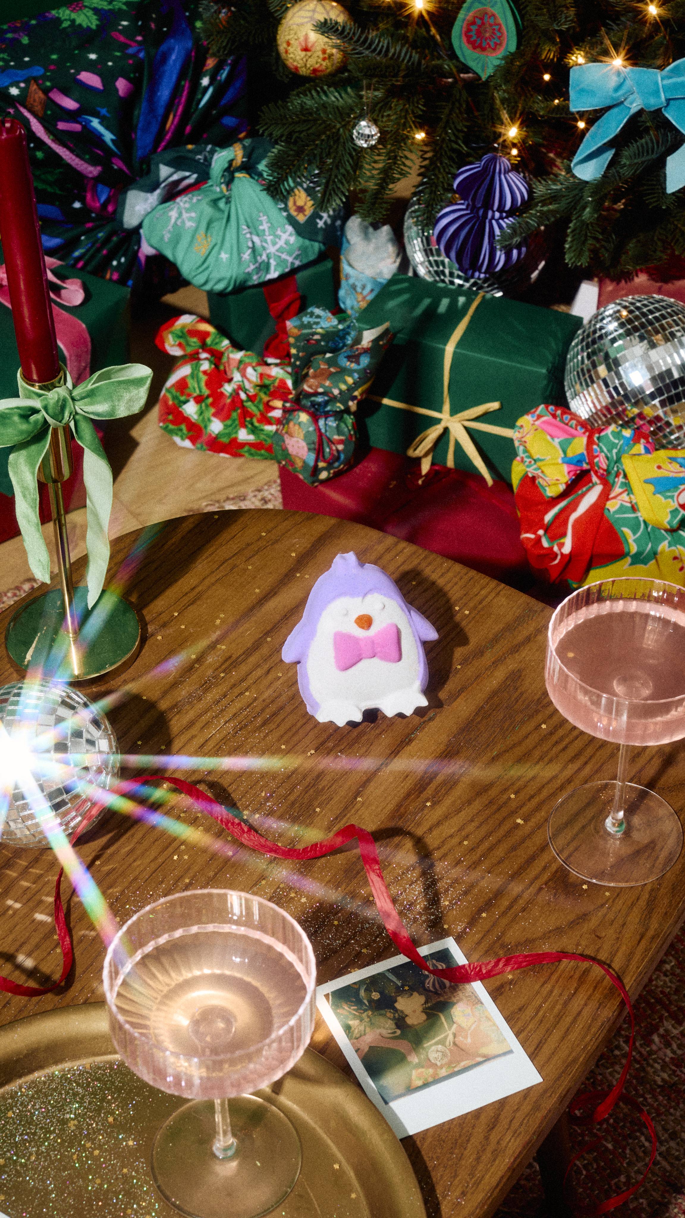 A purple and white penguin bath bomb wearing a pink bow tie sits on a wooden table, surrounded by pink cocktail glasses, red ribbon, and wrapped Christmas gifts under a tree.