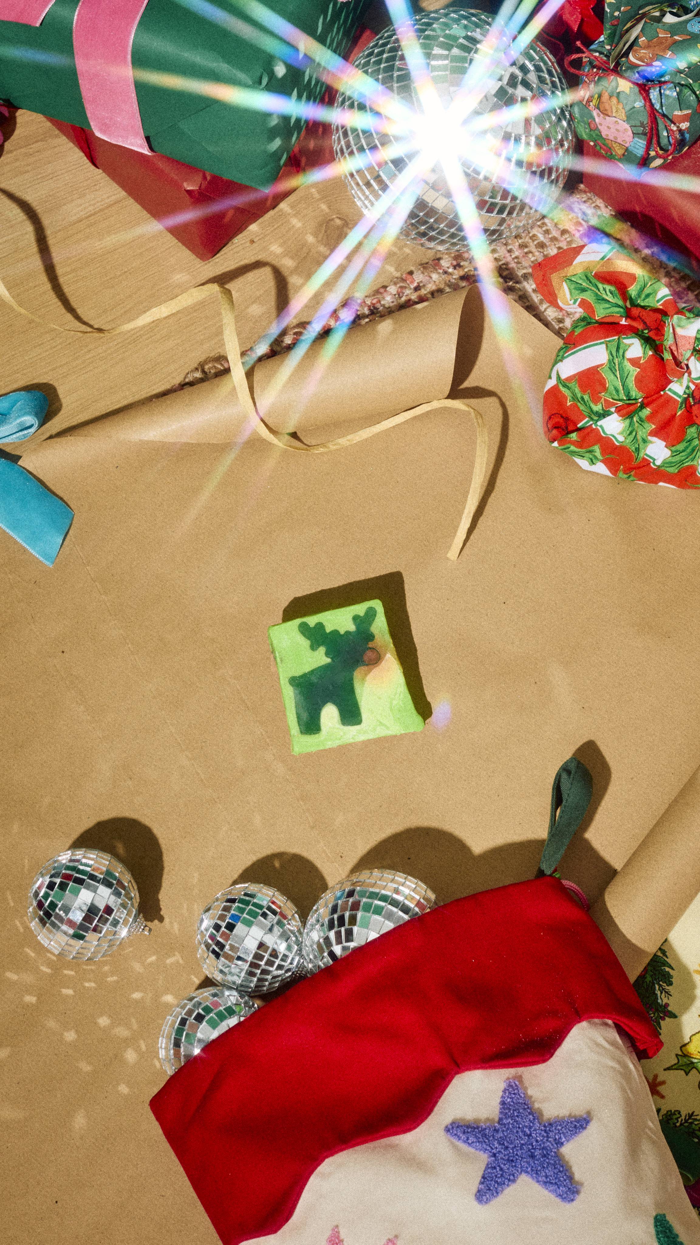 A square lime green soap featuring a dark green reindeer silhouette rests on brown wrapping paper, surrounded by disco ball ornaments, a red Christmas stocking, and festive wrapped presents.