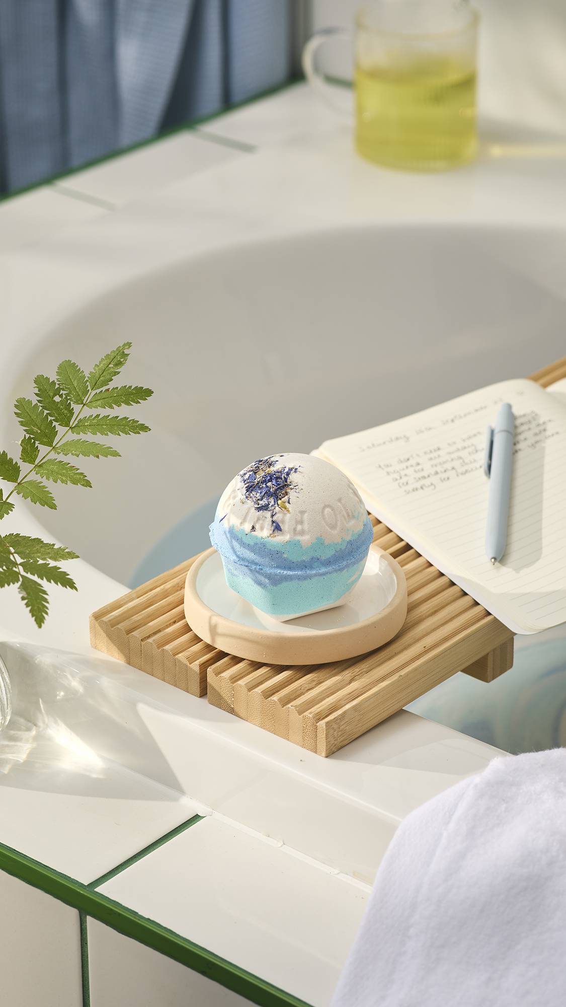 A layered white and blue LUSH "This Too Shall Pass" bath bomb topped with dried blue petals rests on a small dish atop a wooden bath caddy, set alongside an open notebook and pen by a bathtub.