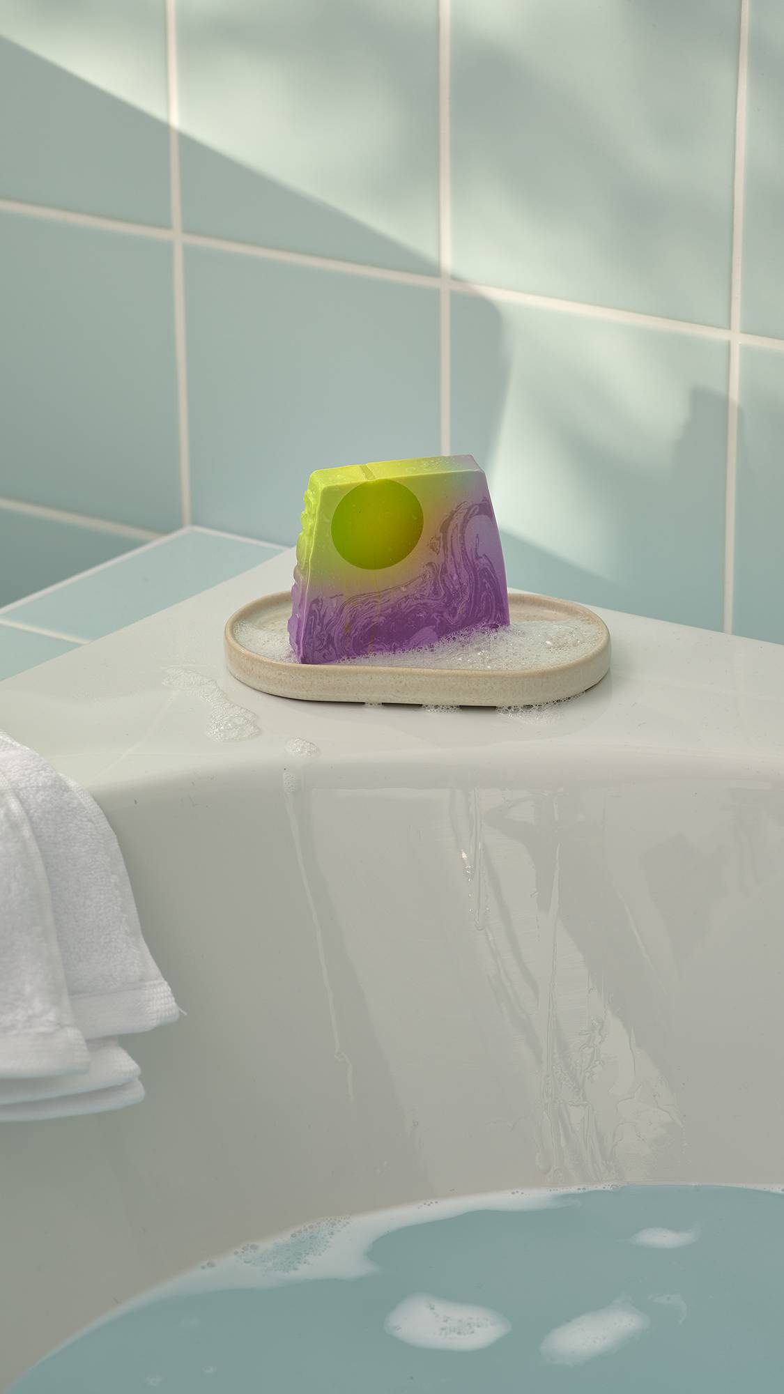 The "To The Moon and Back" green and purple soap can be seen sitting in a small, oval dish on the corner of a white bathtub. The scene is set against mint green wall tiles, with a white towel visible on the left and soapy blue water in the bath.