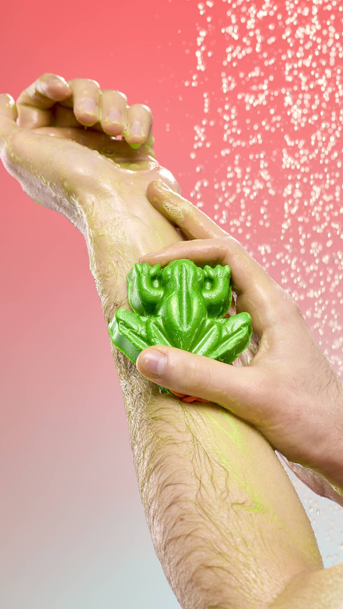  A person lathers their arm with the bright green, frog-shaped Tree Hugger soap under a spray of falling water. The shimmering soap leaves a light green foam on the skin against a pink-to-blue gradient background.