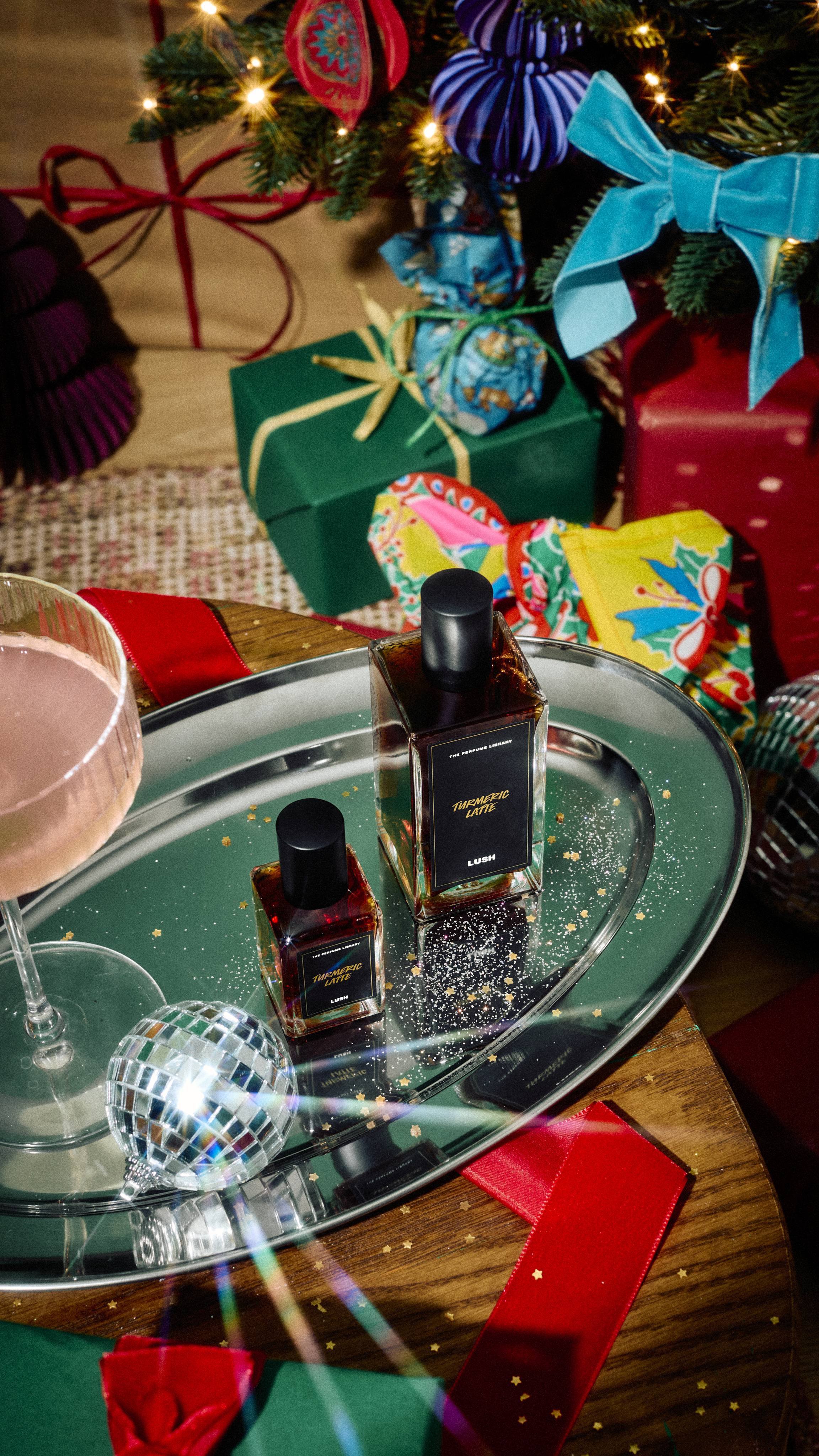 Two glass bottles, one larger and one smaller size, of Lush Turmeric Latte perfume rest on a silver tray scattered with gold glitter, alongside a cocktail glass and mini disco ball, set against a festive background of wrapped gifts.
