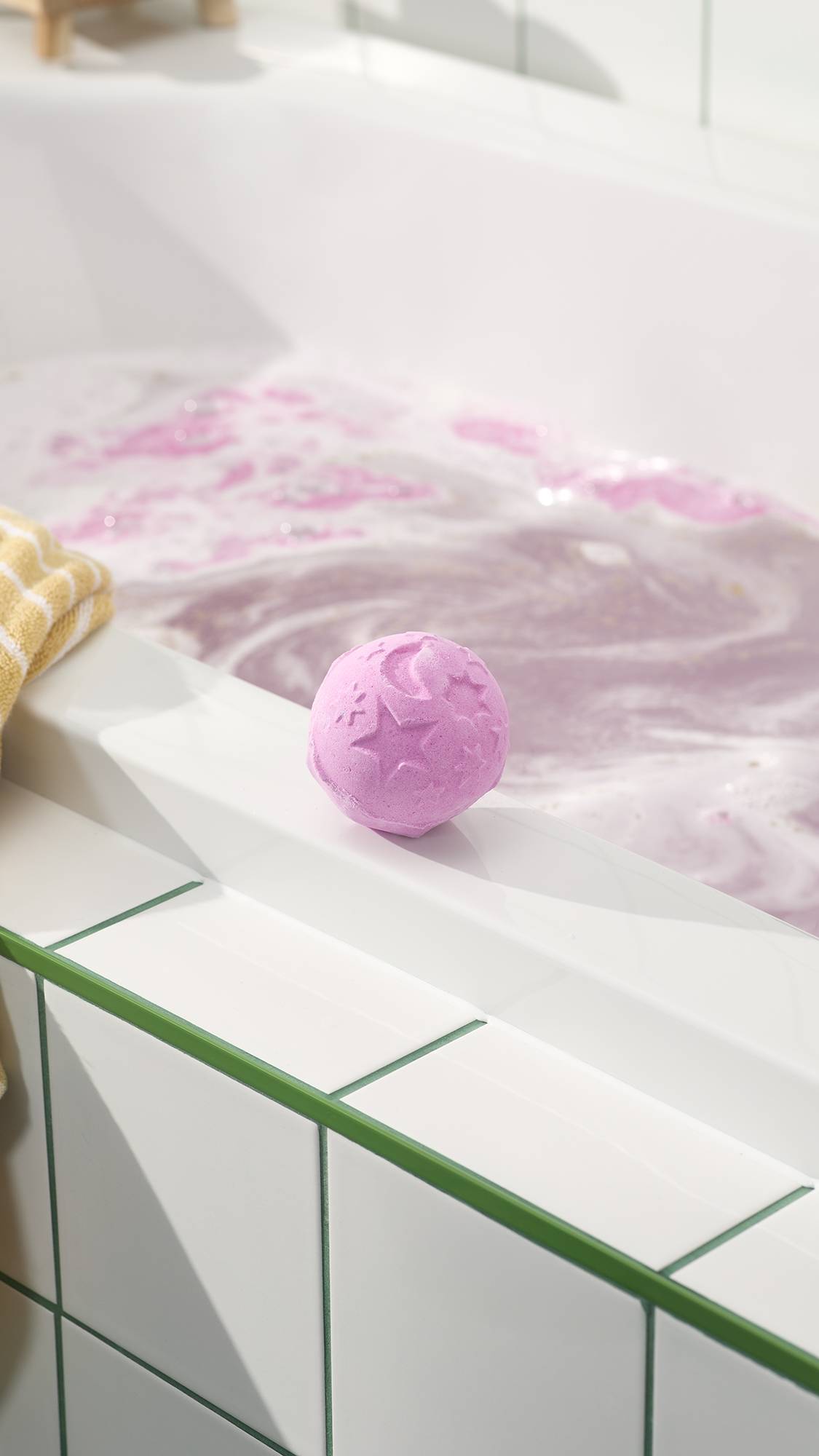 The Twilight Bath Bomb sits on the clean, white edge of a bathtub, overlooking water already filled with swirling lavender and pink foam. This celestial-themed bath bomb is a soft purple sphere decorated with embossed stars and a crescent moon.