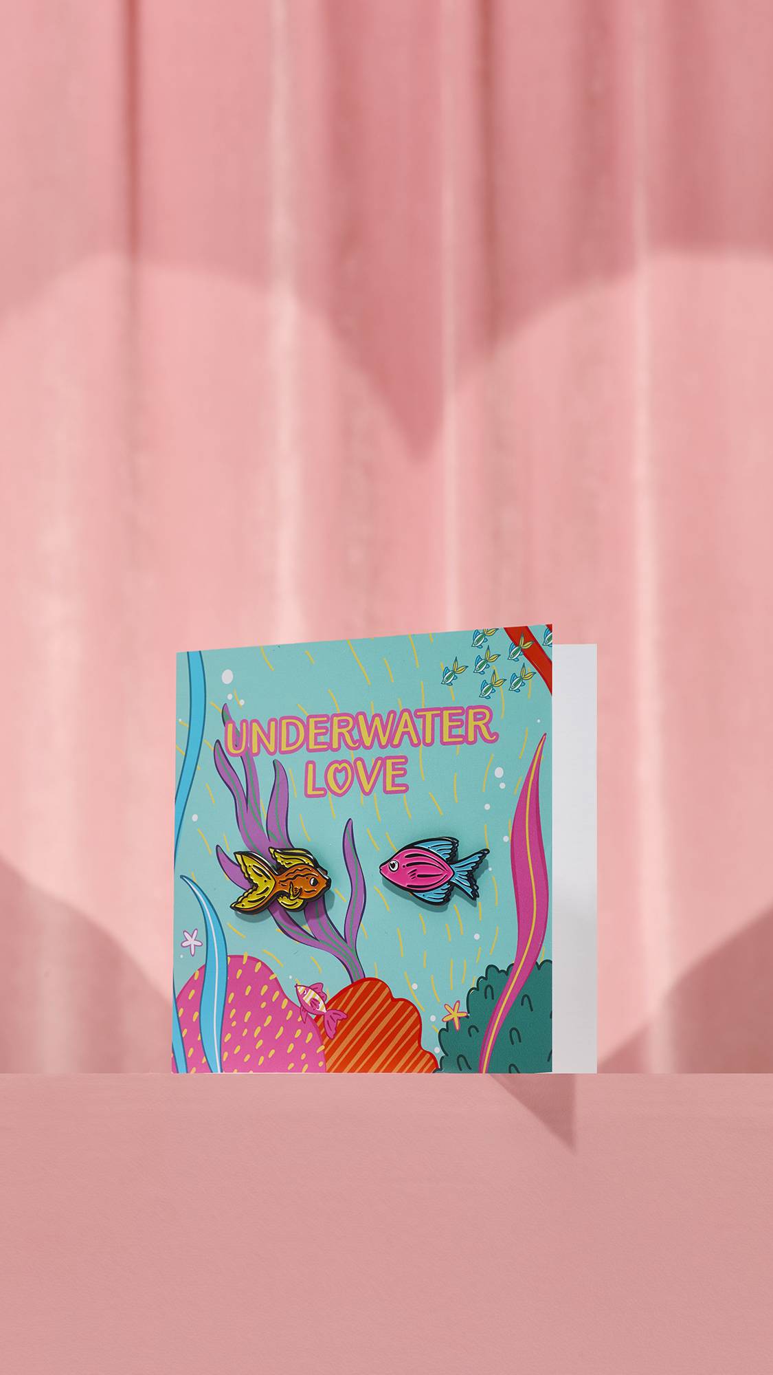 The Underwater Love card features a light blue aquatic scene with pink "UNDERWATER LOVE" text. Two fish pins—one gold, one pink—are attached to the front amongst colorful coral illustrations against a pink background.