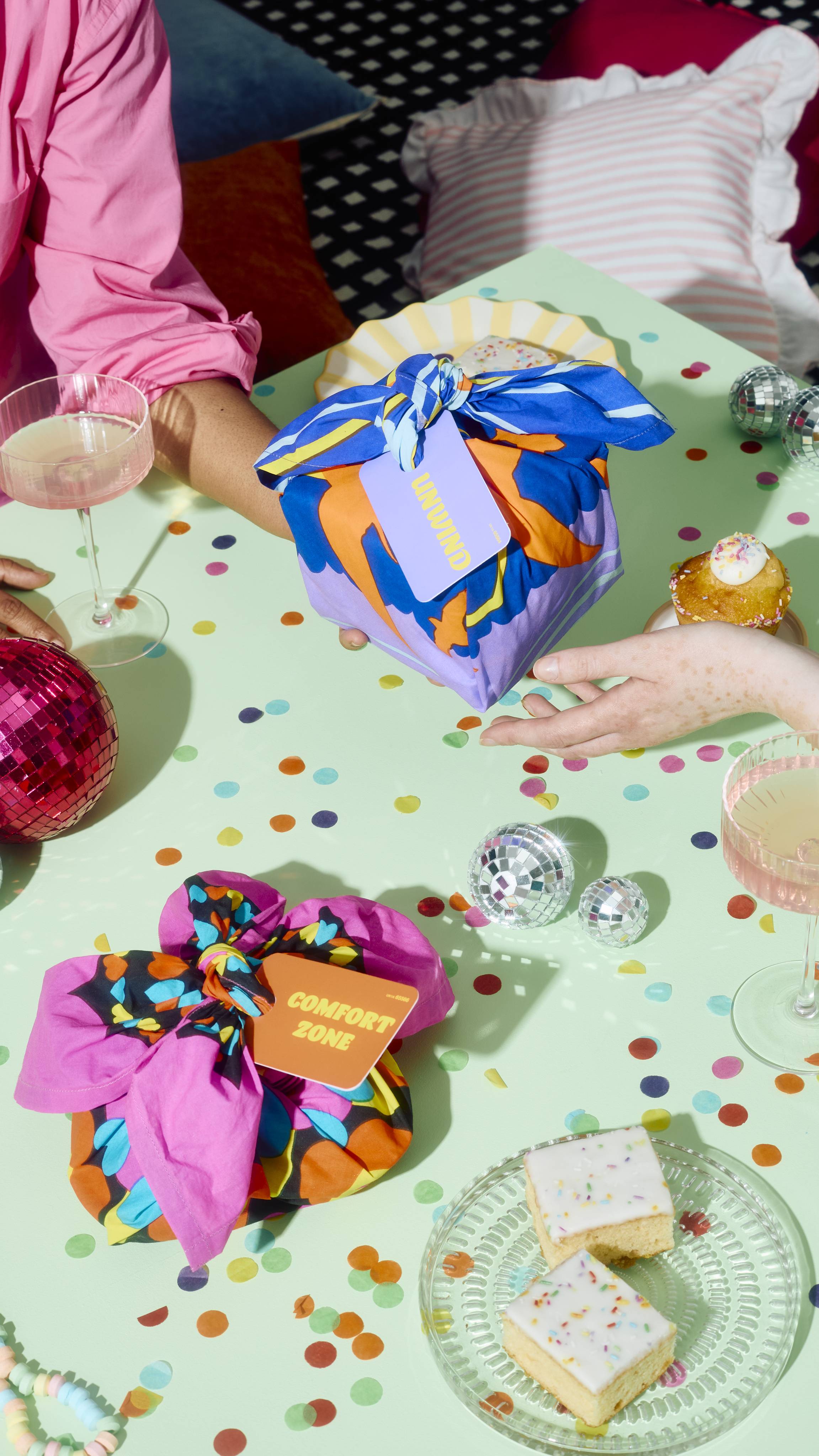 Two hands exchange a knot-wrapped "UNWIND" gift across a pastel green party table scattered with confetti, mini disco balls, iced cakes, and a pink gift tagged "COMFORT ZONE".