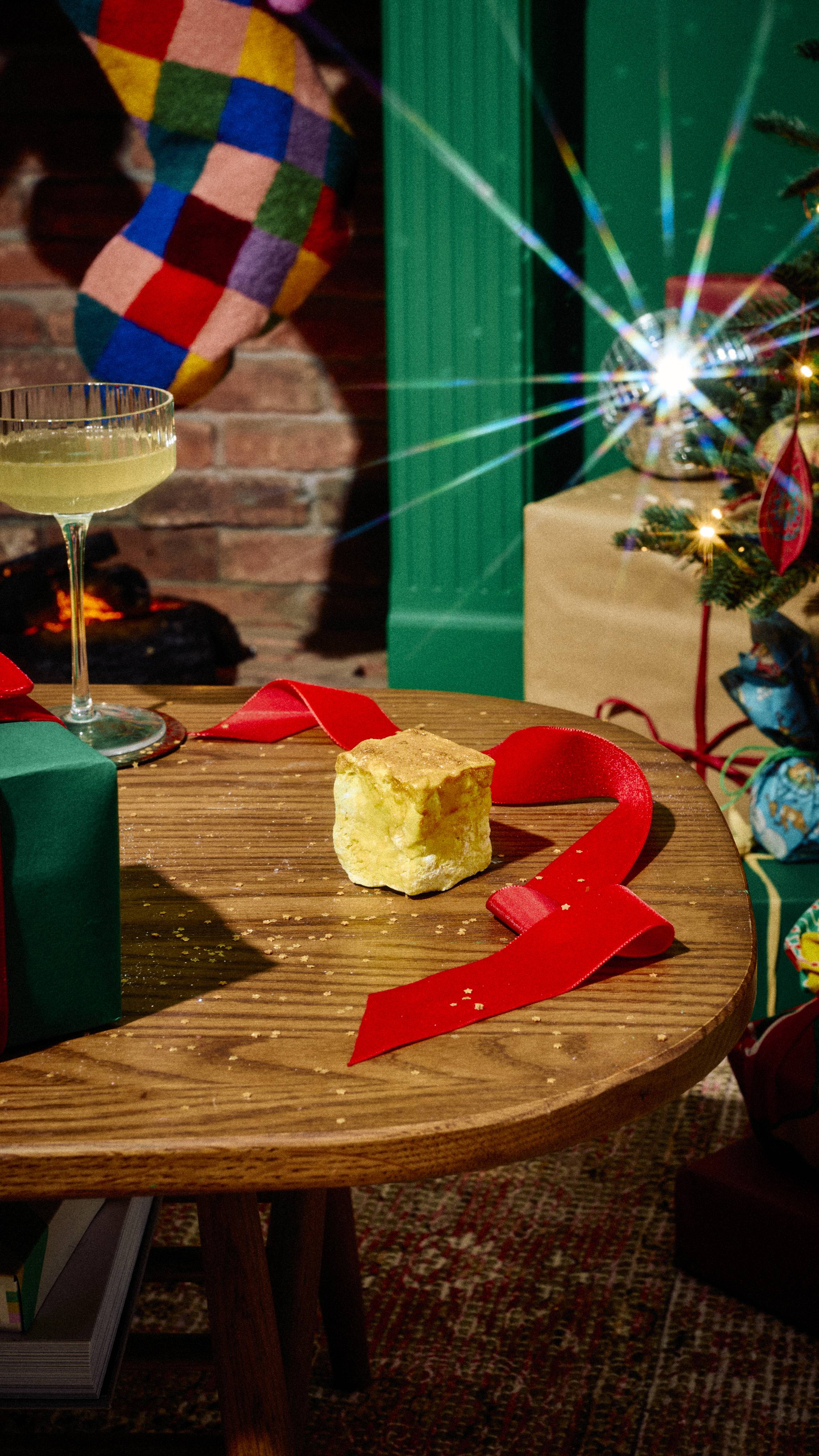 A small, textured yellow cube of Lush "Vanilla Delight" soap rests on a wooden table beside red ribbon, a cocktail glass, and wrapped presents, set against a festive background with a colourful stocking.