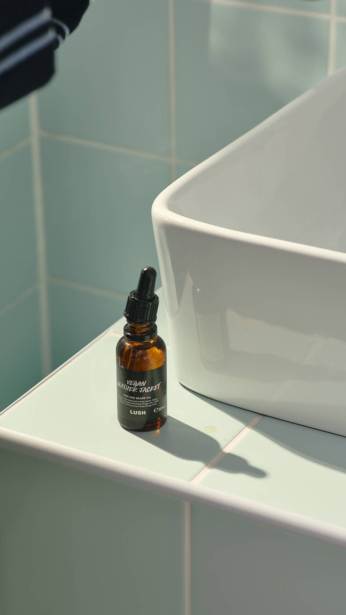 A small, amber glass bottle of "Vegan Leather Jacket" hair and beard oil by Lush sits on a pale green tiled ledge next to a white ceramic sink. The bottle features a black label and a pipette dropper lid, casting a soft shadow on the tiles.