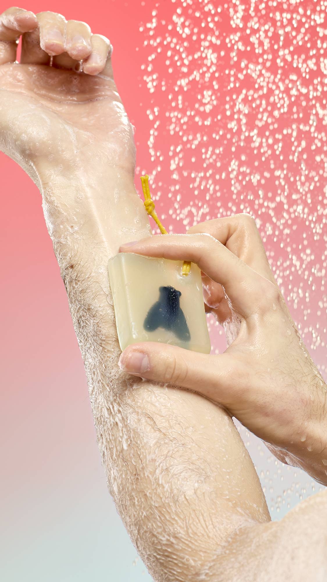 A person lathers their arm with Waiter! There's A Fly In My Soap under a spray of water. The translucent yellow bar features a fly silhouette and a yellow cord. Set against a pink-to-blue gradient background.