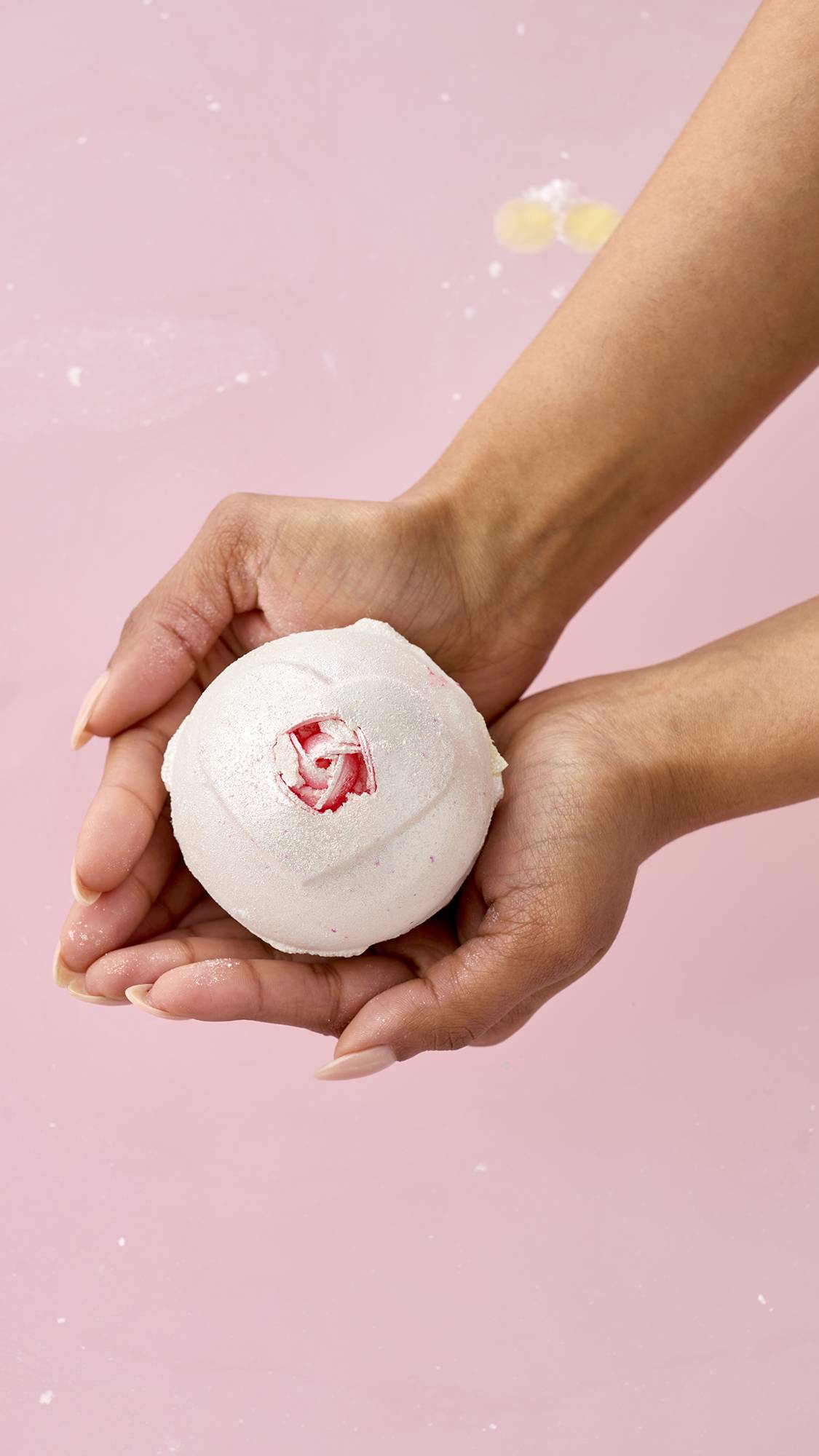 A pair of hands holds the pearlescent white Wedding bath bomb over pink bathwater. The sphere features an embossed heart shape with a small, realistic pink rosebud at its center.