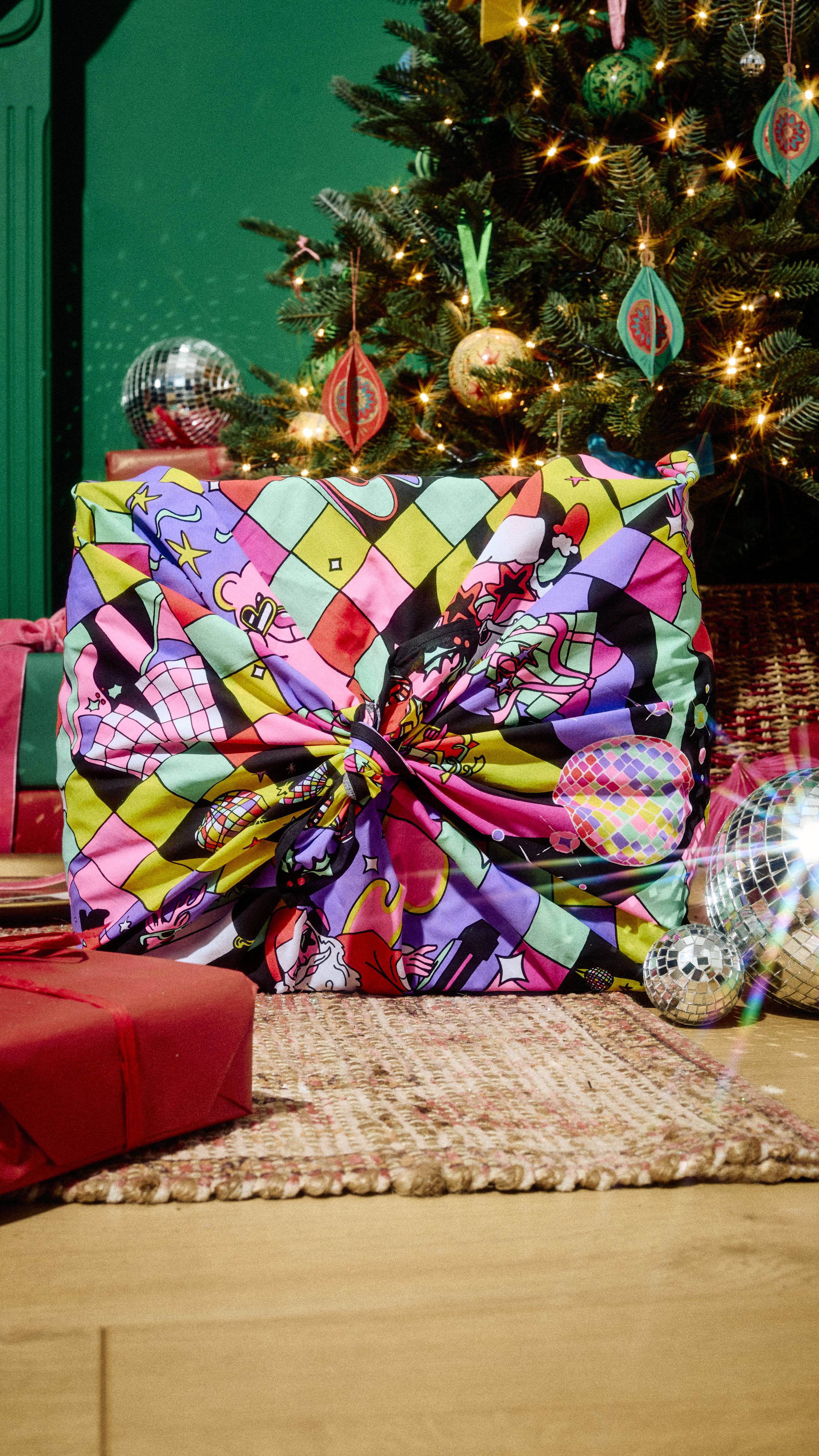 A present wrapped in a colourful, festive disco-patterned Lush fabric Knot Wrap, tied in a knot on top, resting on a patterned rug beside a decorated Christmas tree and shiny disco balls.