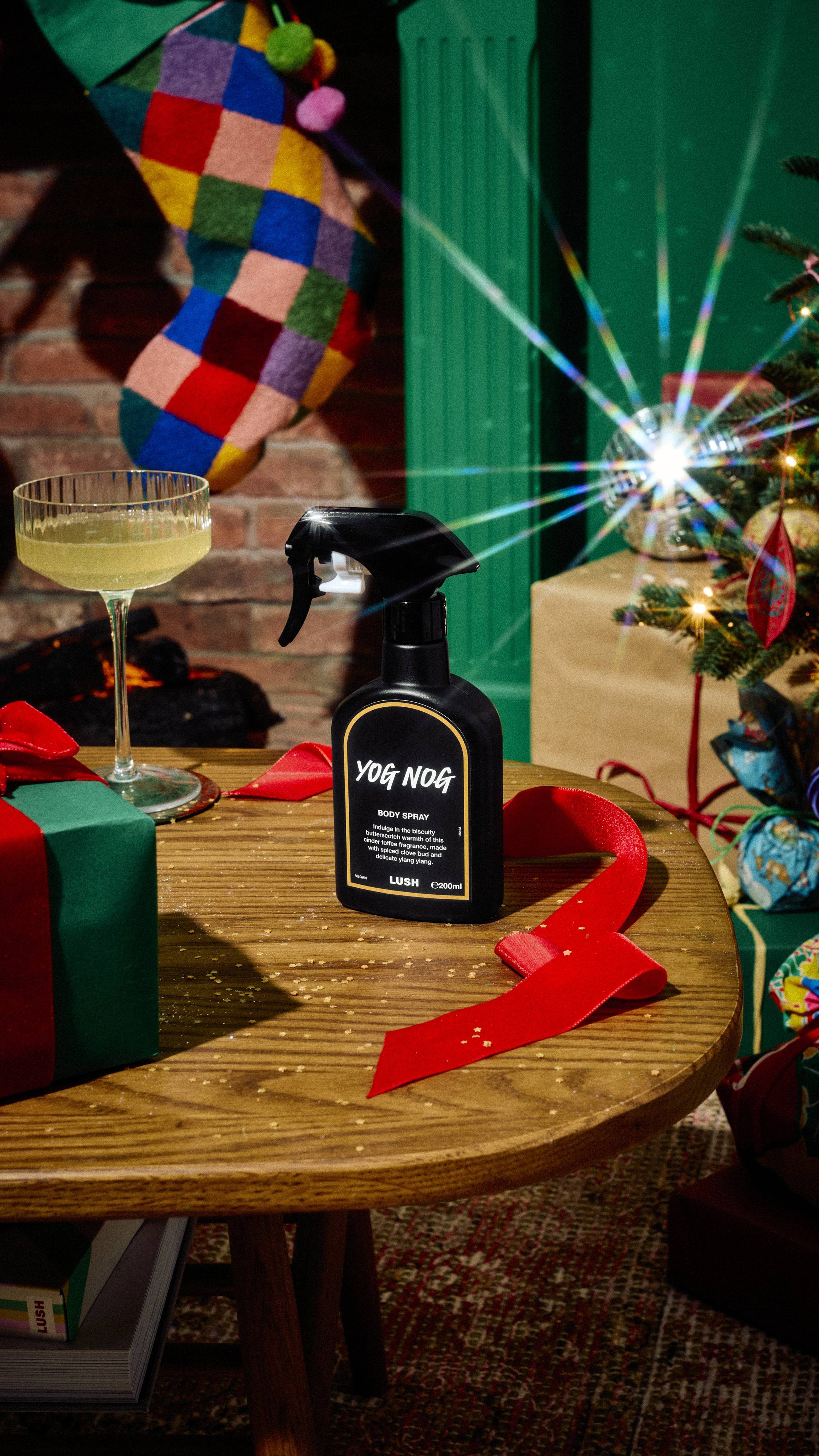 A black spray bottle of Lush Yog Nog body spray stands on a wooden table beside red ribbon, a cocktail glass, and a wrapped gift, set in a festive room with a Christmas tree and colourful stocking.