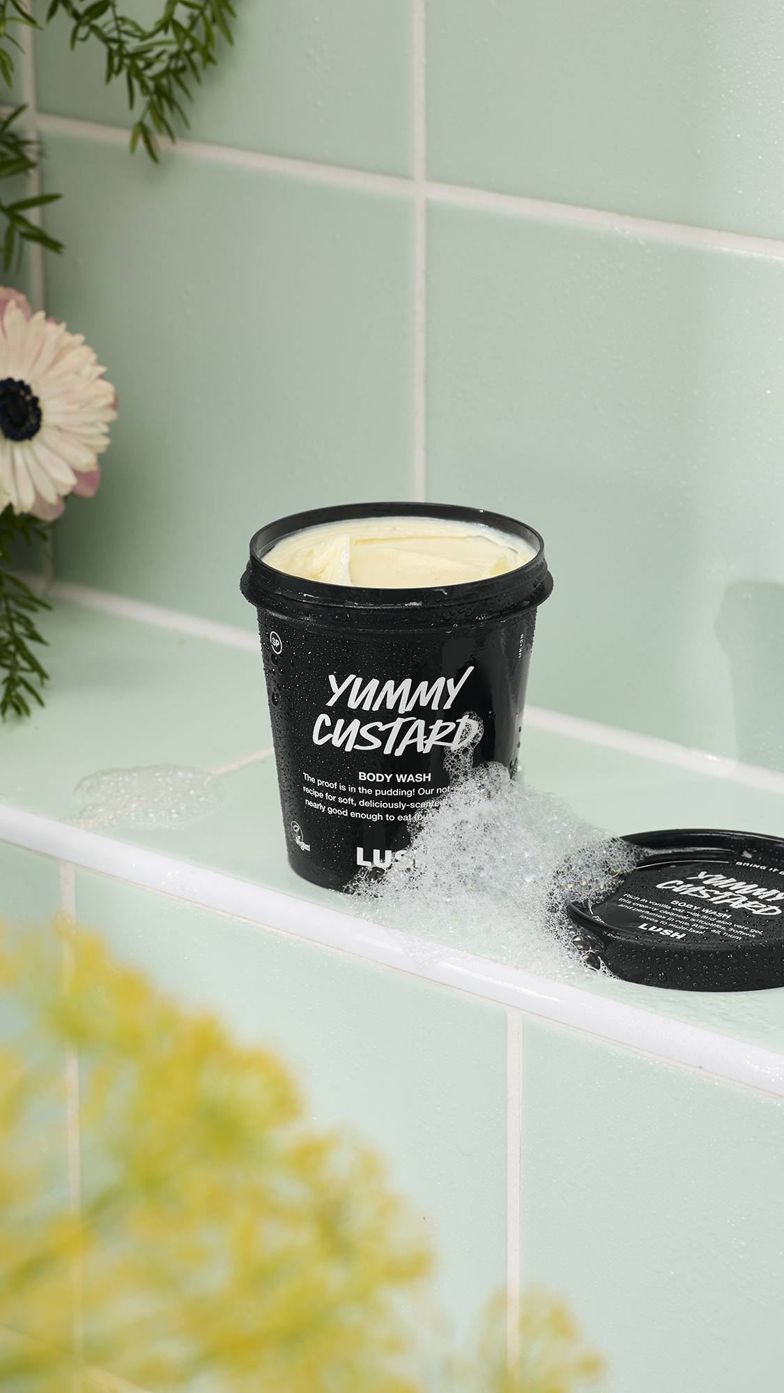 An open black pot of "Yummy Custard" body wash sits on a white tiled ledge, filled with a thick, creamy yellow product. White soapy bubbles cluster around the base of the tub, while mint green tiles and floral accents create a fresh bathroom setting.