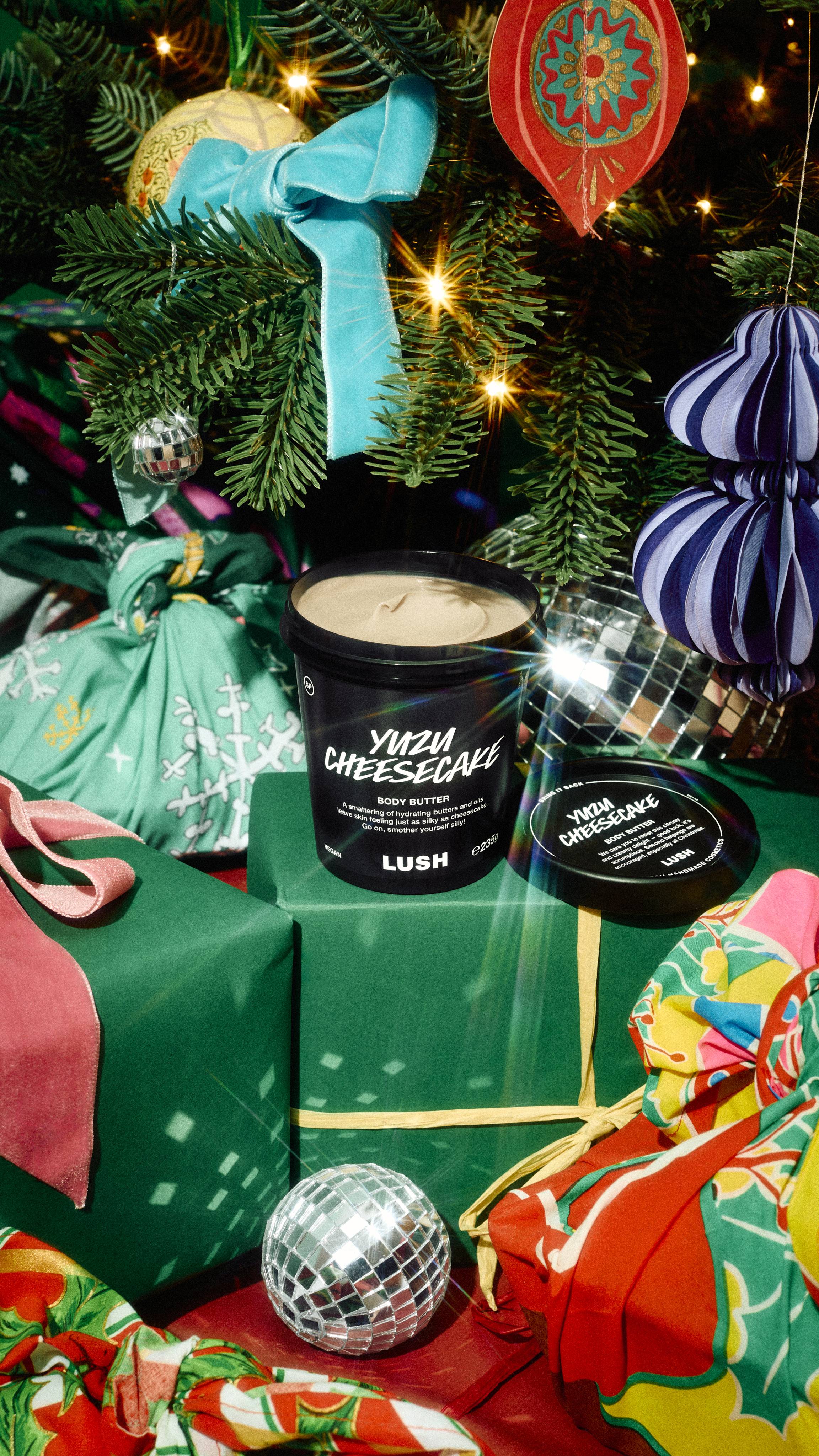 An open black tub of Lush Yuzu Cheesecake body butter sits atop green wrapped presents next to mini disco balls under a decorated Christmas tree.