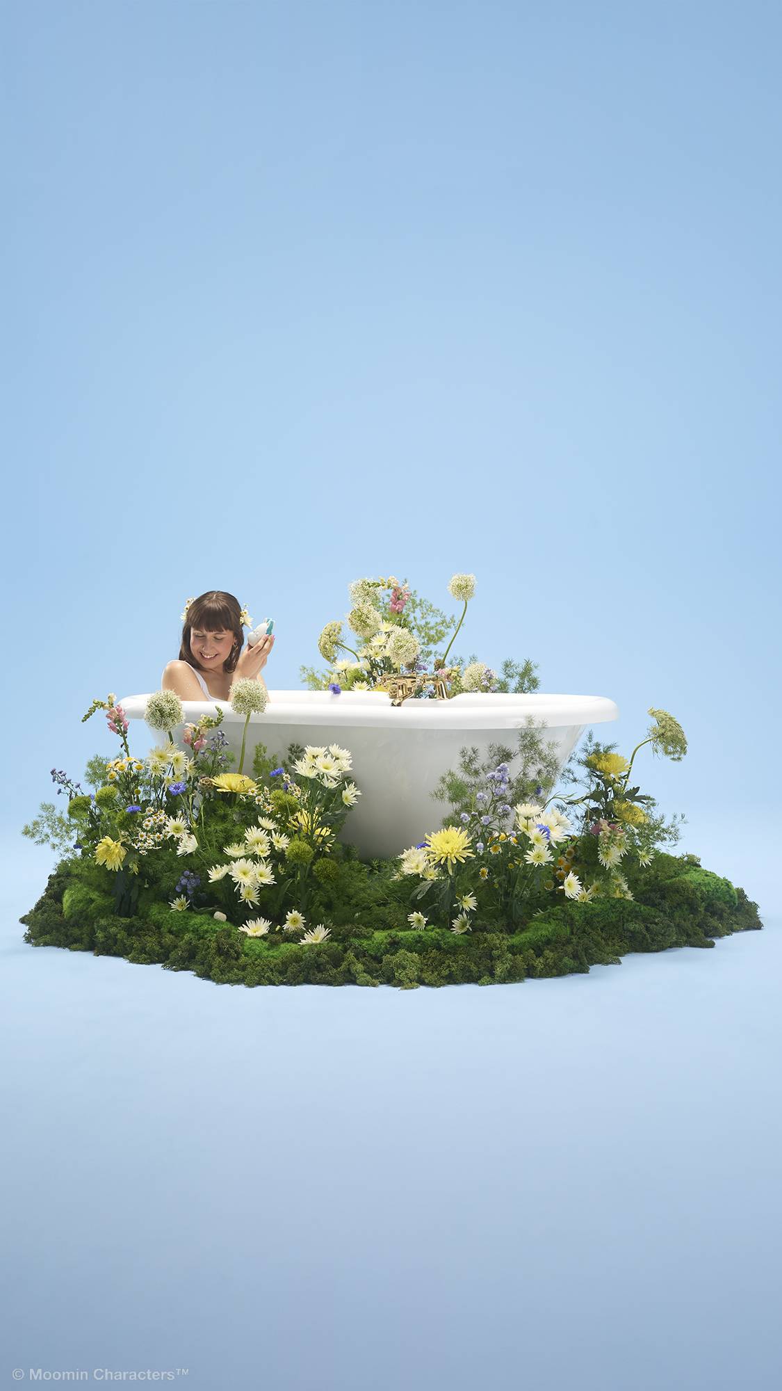 A person smiles whilst sitting inside a white bathtub, holding a small bath bomb. The tub is placed on a patch of lush green moss overflowing with yellow and white wildflowers, set against a plain light blue background.