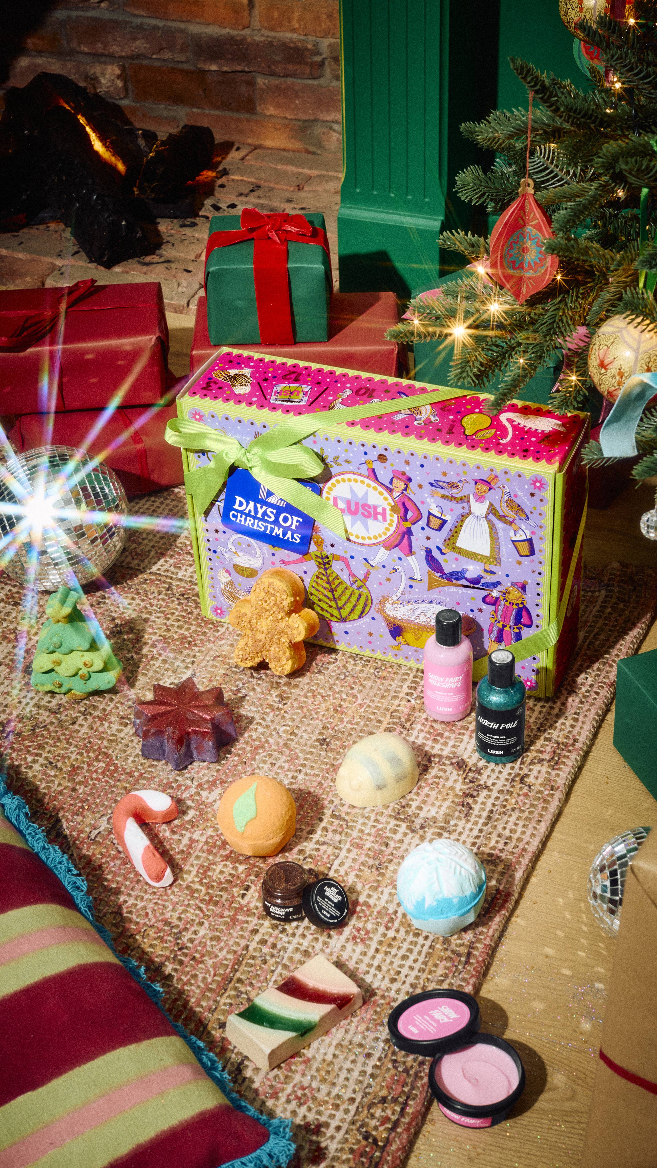 The Lush 12 Days of Christmas gift box sits on a patterned rug surrounded by 12 products, including bath bombs, shower gels, soap, lip scrub, and bath treats, in a festive room setting by a fireplace and a decorated Christmas tree.