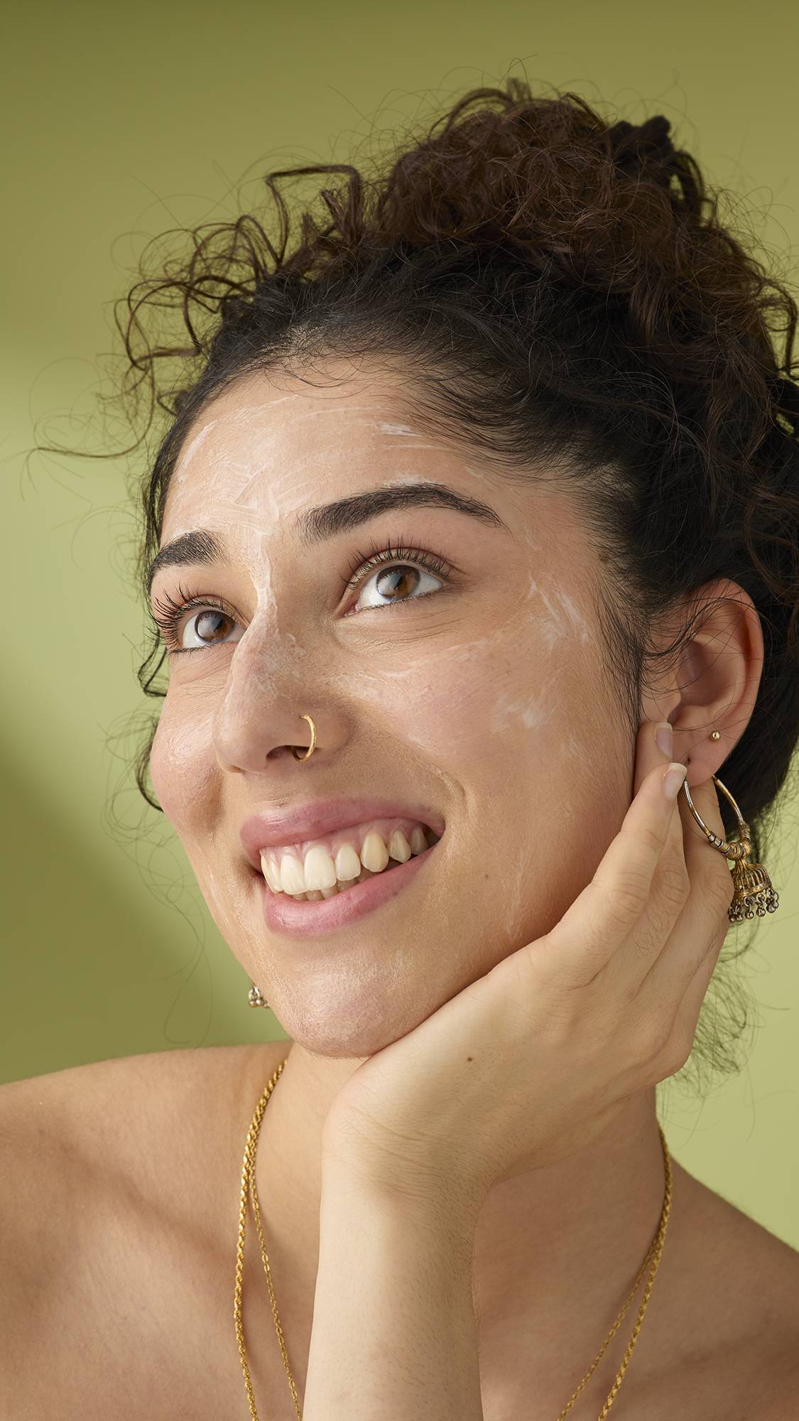 A person with curly hair and a subtle nose ring smiles brightly while using a white pad to apply the 9 to 5 cleanser to their cheek