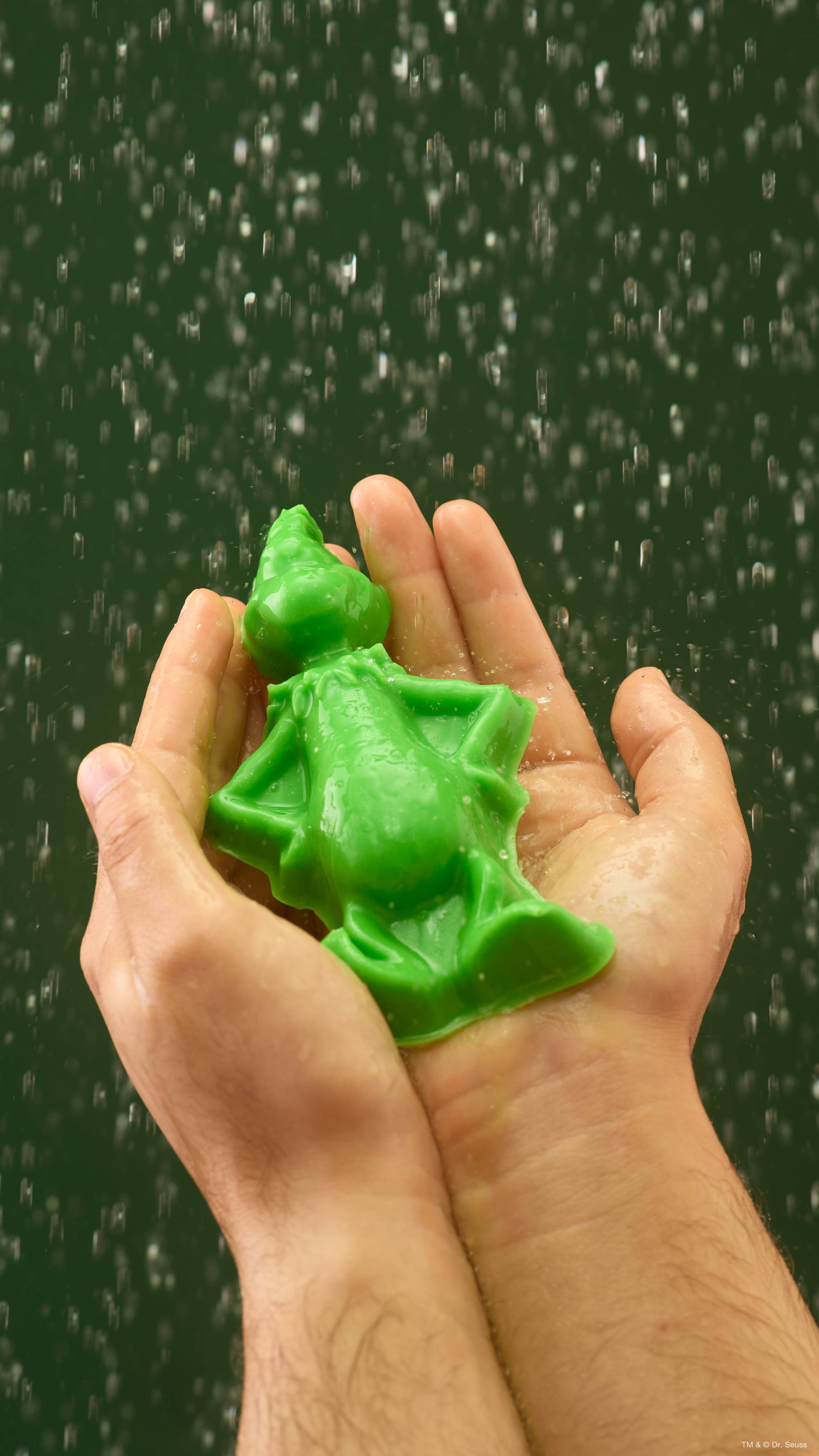 Two hands cupping a bright green Lush shower gummy shaped like The Grinch under streaming water droplets, set against a dark green backdrop.  