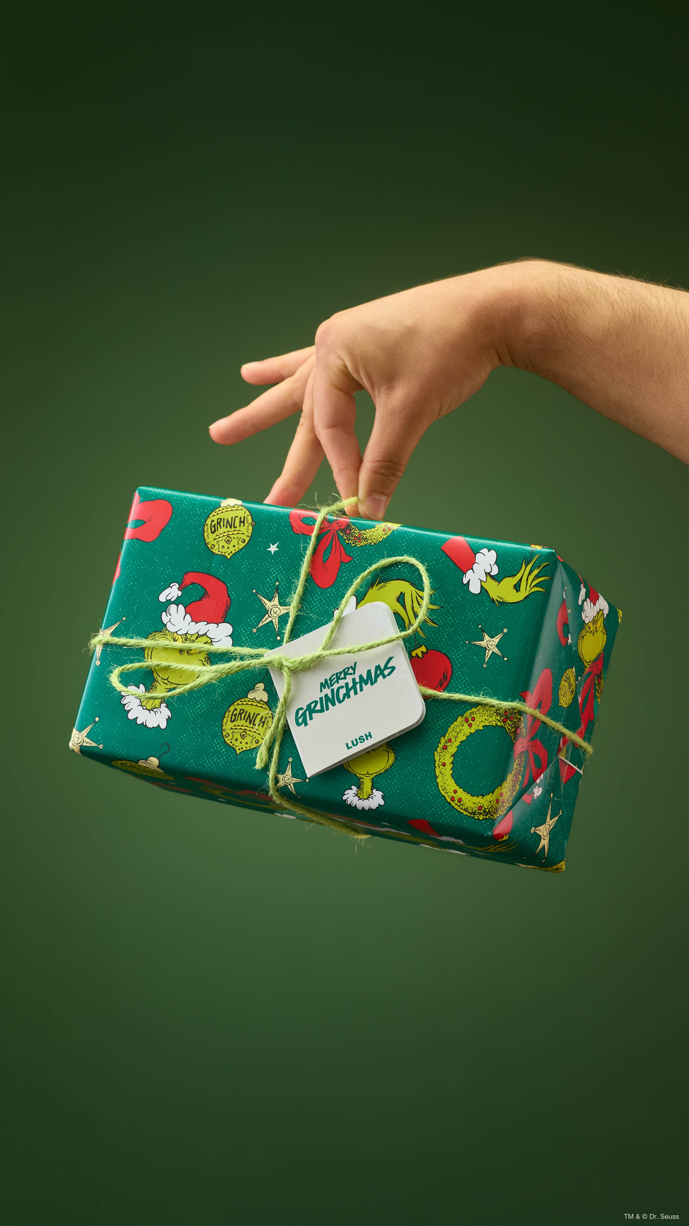 A hand lifts a Lush 'Merry Grinchmas' gift box by its green string bow. The rectangular box is wrapped in festive Grinch-themed paper with a white gift tag reading 'Merry Grinchmas', set against a solid dark green background.  