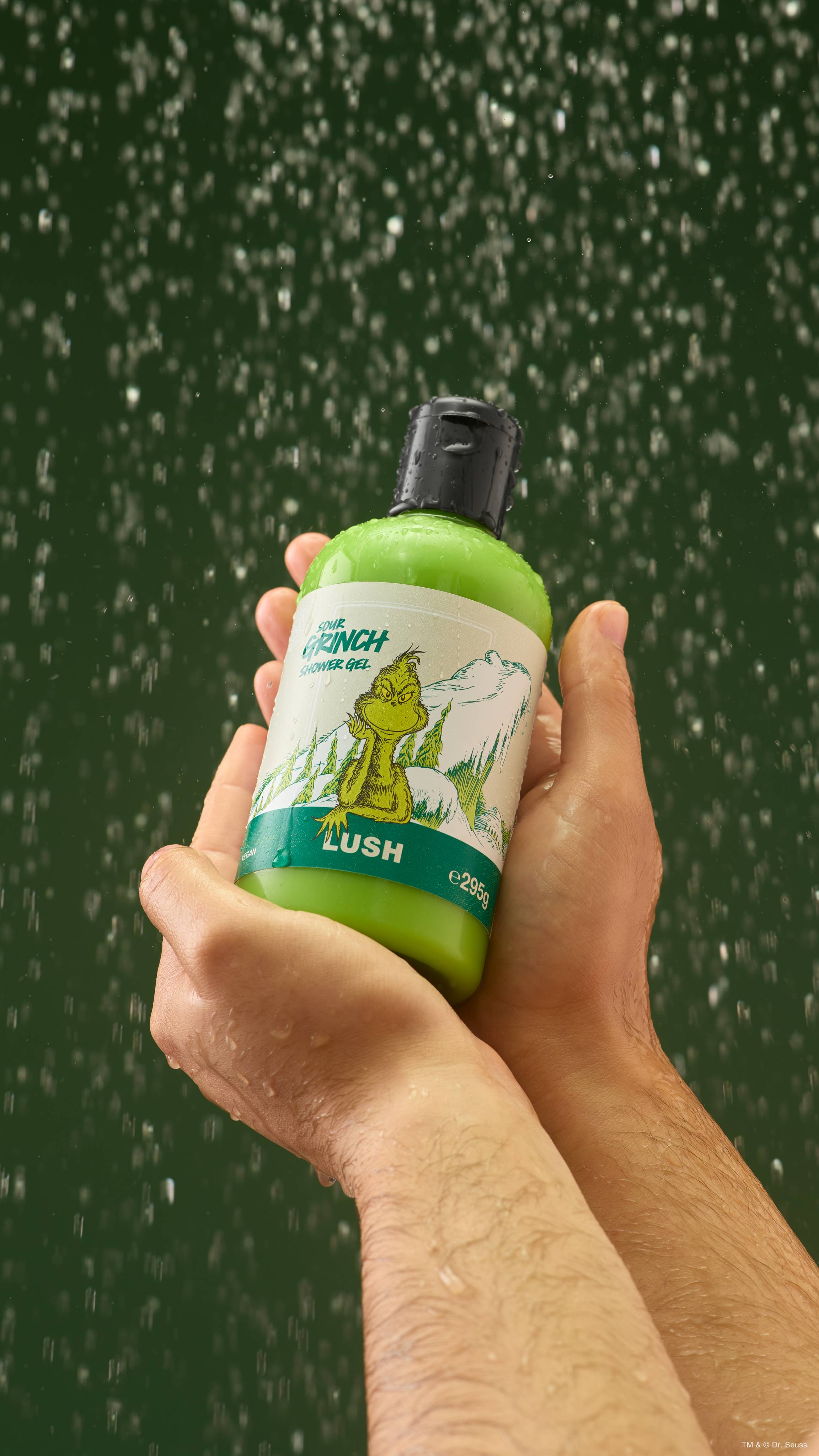 Two hands cupping a bottle of Lush 'Sour Grinch' bright green shower gel under falling water droplets, set against a dark green backdrop.  
