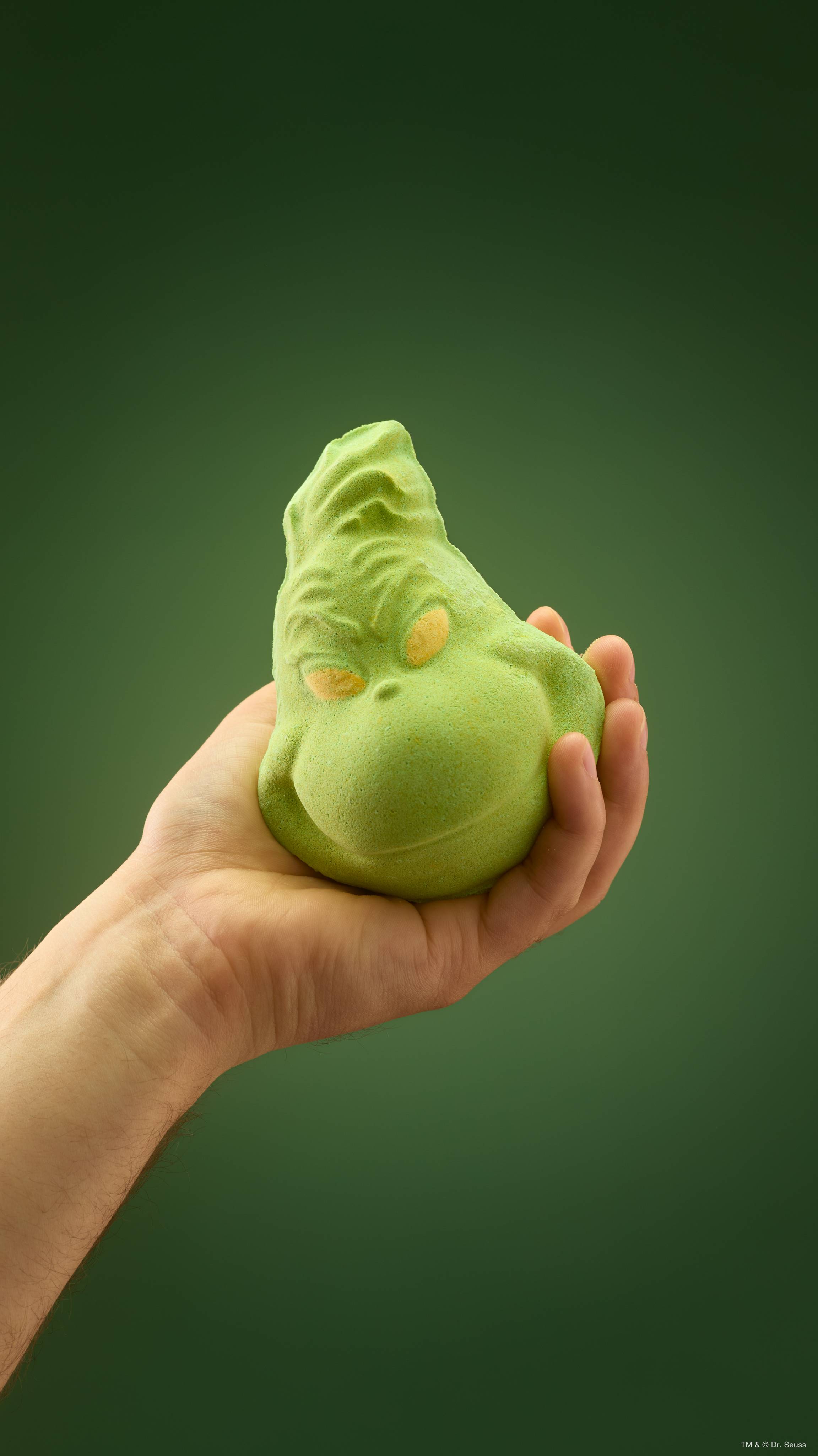 A hand holds a lime-green Lush bath bomb shaped like a mischievous "Grinch" face with yellow eyes and a smirk, held against a plain green background.  