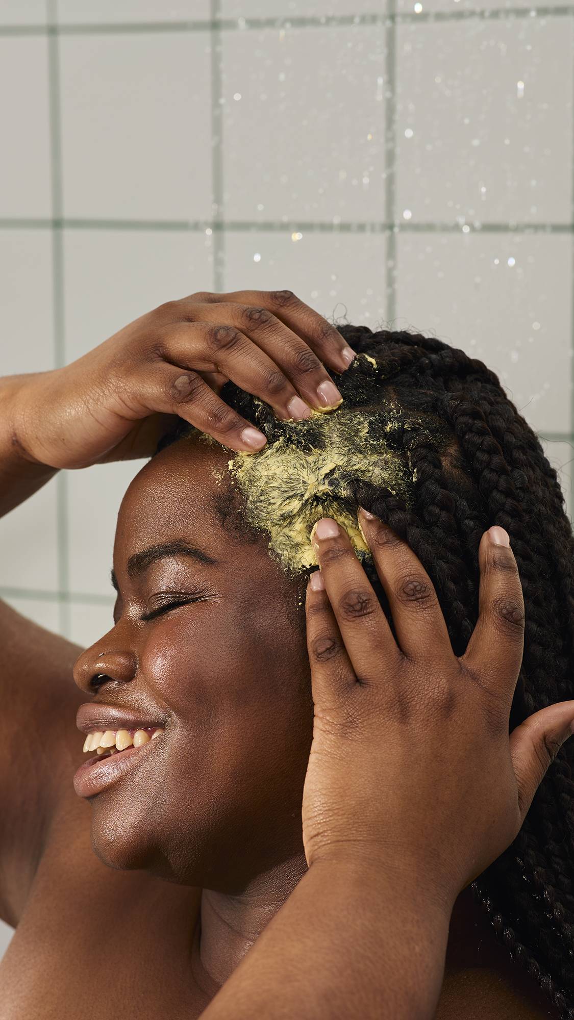 A close-up of a person with long dark braids smiling as they massage a mustard-yellow "Ahaa" treatment into their scalp. In the background, water falls against white square tiles with green grout