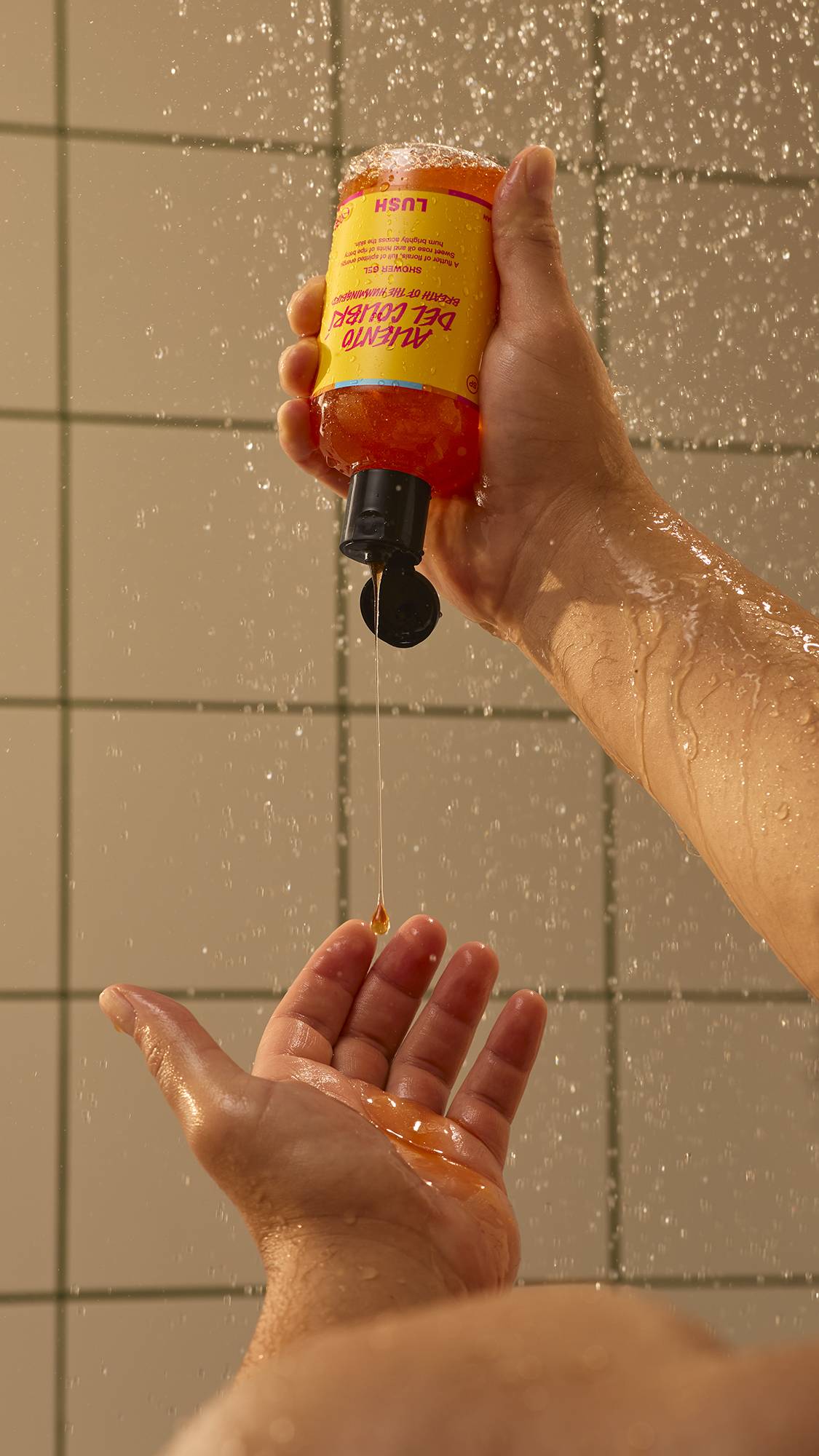 A person squeezes orange Lush Aliento Del Colibrí shower gel from an inverted bottle into their palm under a warm spray of water, against a tiled bathroom wall.