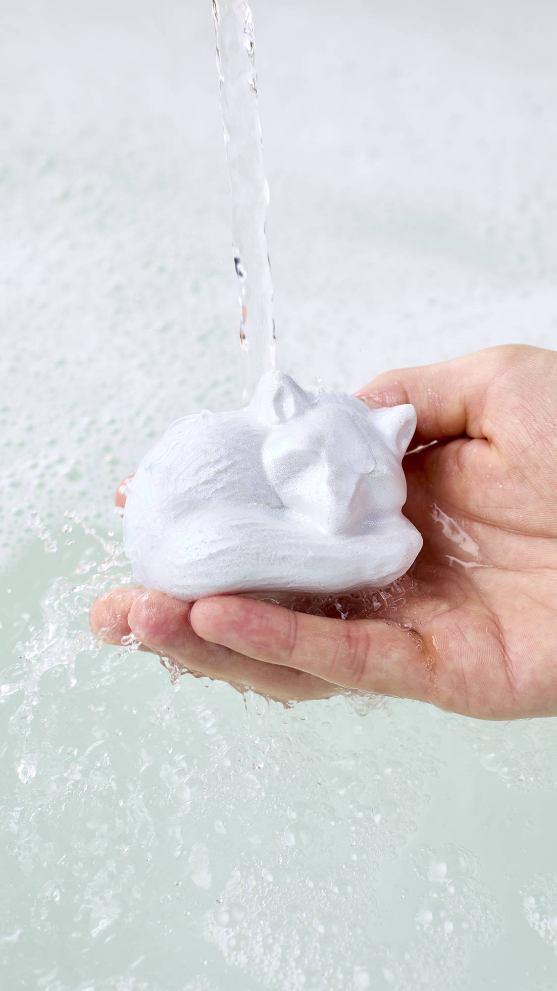 A hand holds a white, sleeping fox-shaped bubble bar under a gentle stream of running tap water, splashing over milky white bathwater covered in light, airy bubbles.