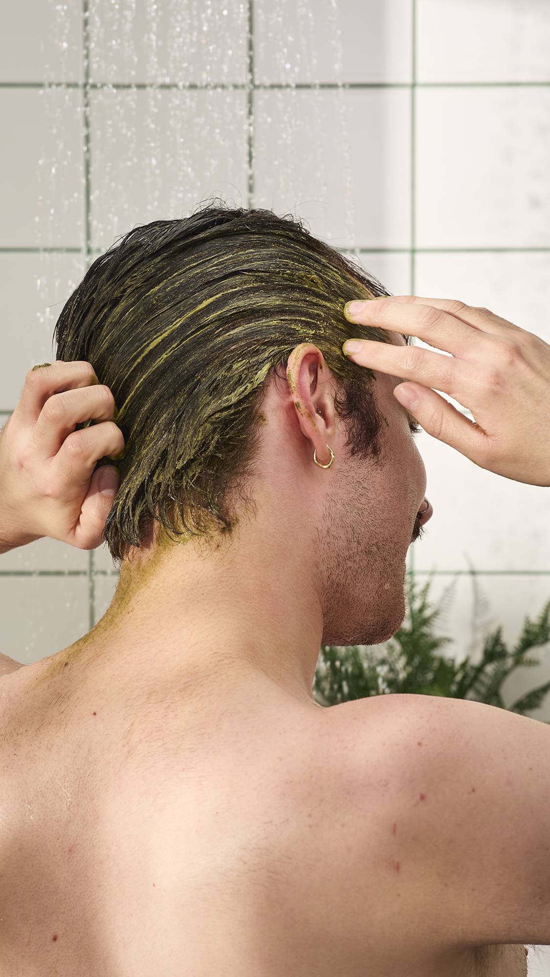 A close-up shows a person from behind, massaging a thick, olive-green "Babylon" scalp treatment into their short hair in a shower. Water falls in the background against white tiles with green grout. 