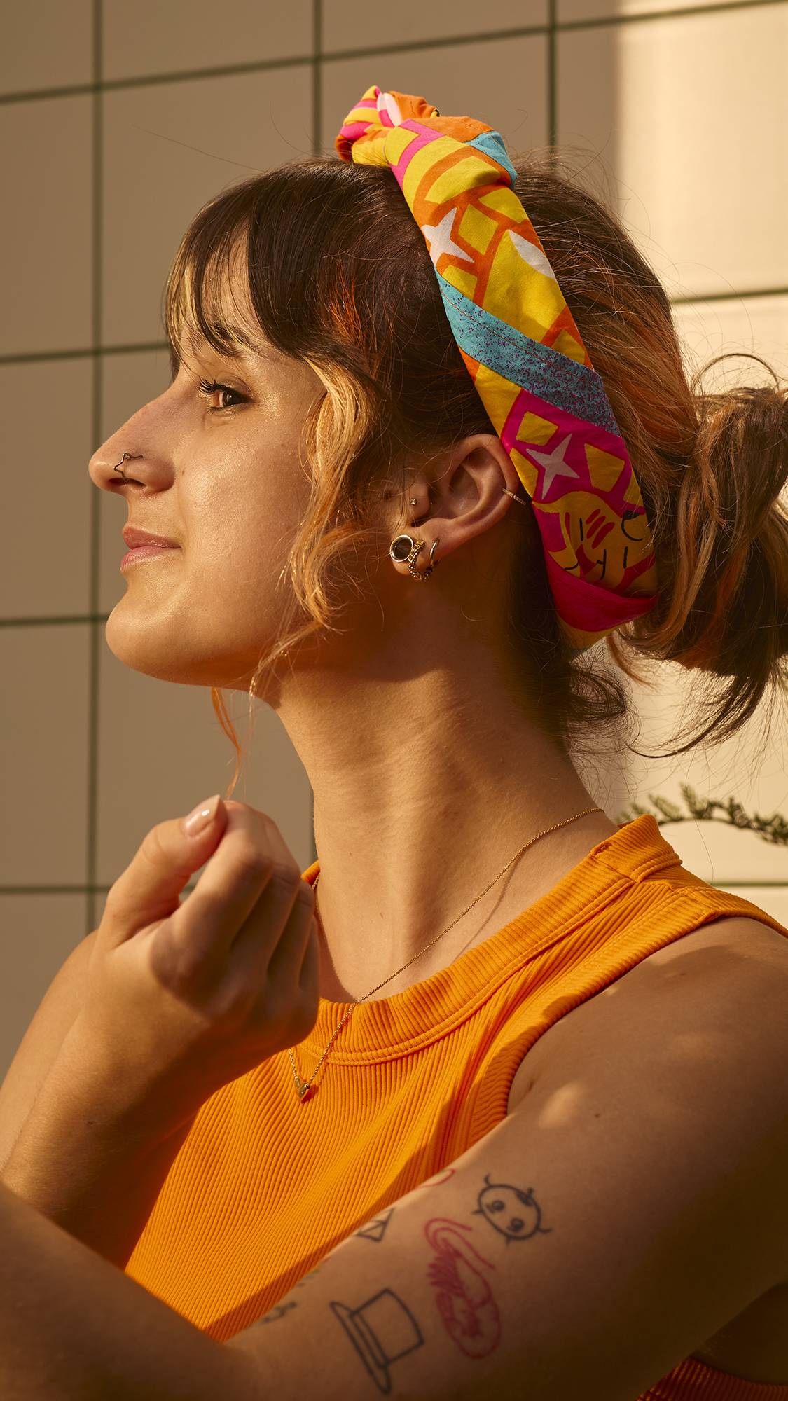 The image shows the side profile of a model wearing a vibrant yellow tank top as they fashion the Lush Bailale Calaverita Knot Wrap into a trendy headscarf, set against a tiled wall in warm sunlight