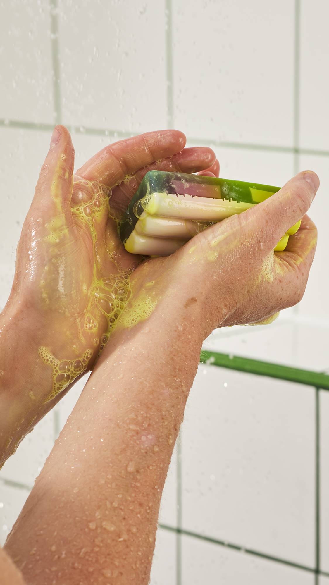 Hands lather a rectangular green and pastel soap bar in the shower, creating light yellowish-green bubbles across wet palms and arms against a white tiled wall.