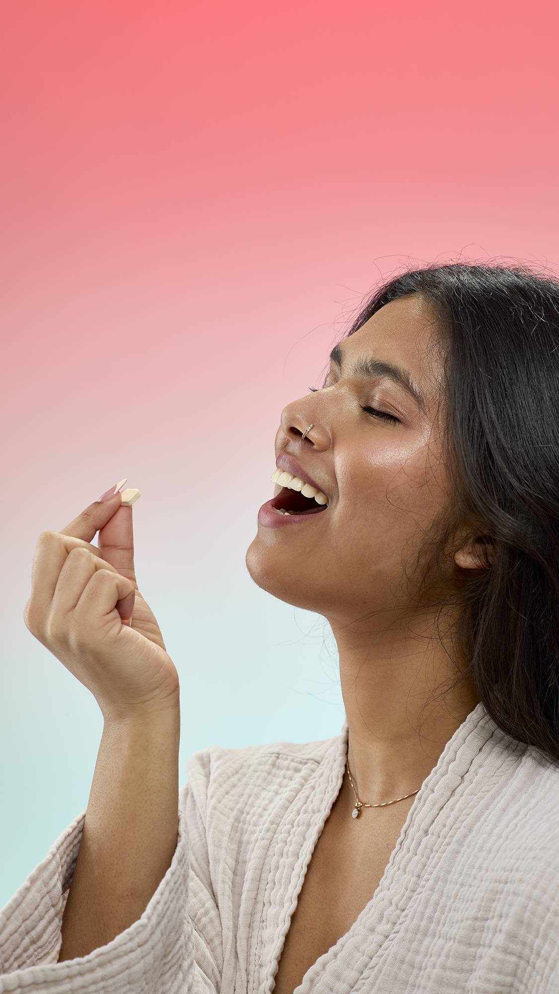 A person with long dark hair and a nose ring smiles with their eyes closed, holding a small, diamond-shaped Banana Bread mouthwash tab toward their open mouth.