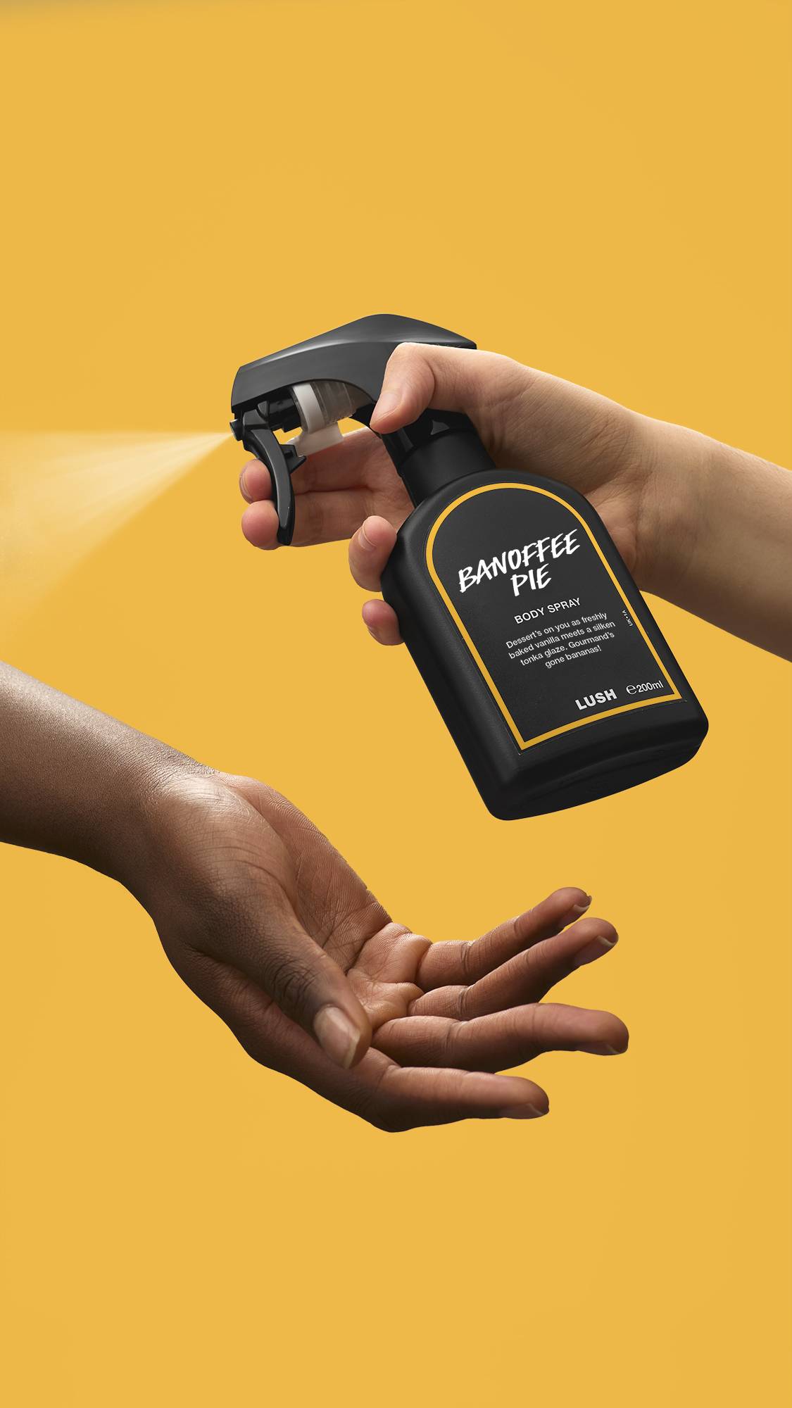 A person's hand presses the nozzle of a black Lush 'Banoffee Pie' body spray bottle, releasing a fine mist onto another person's open palm. The action is set against a solid, vibrant yellow background. 