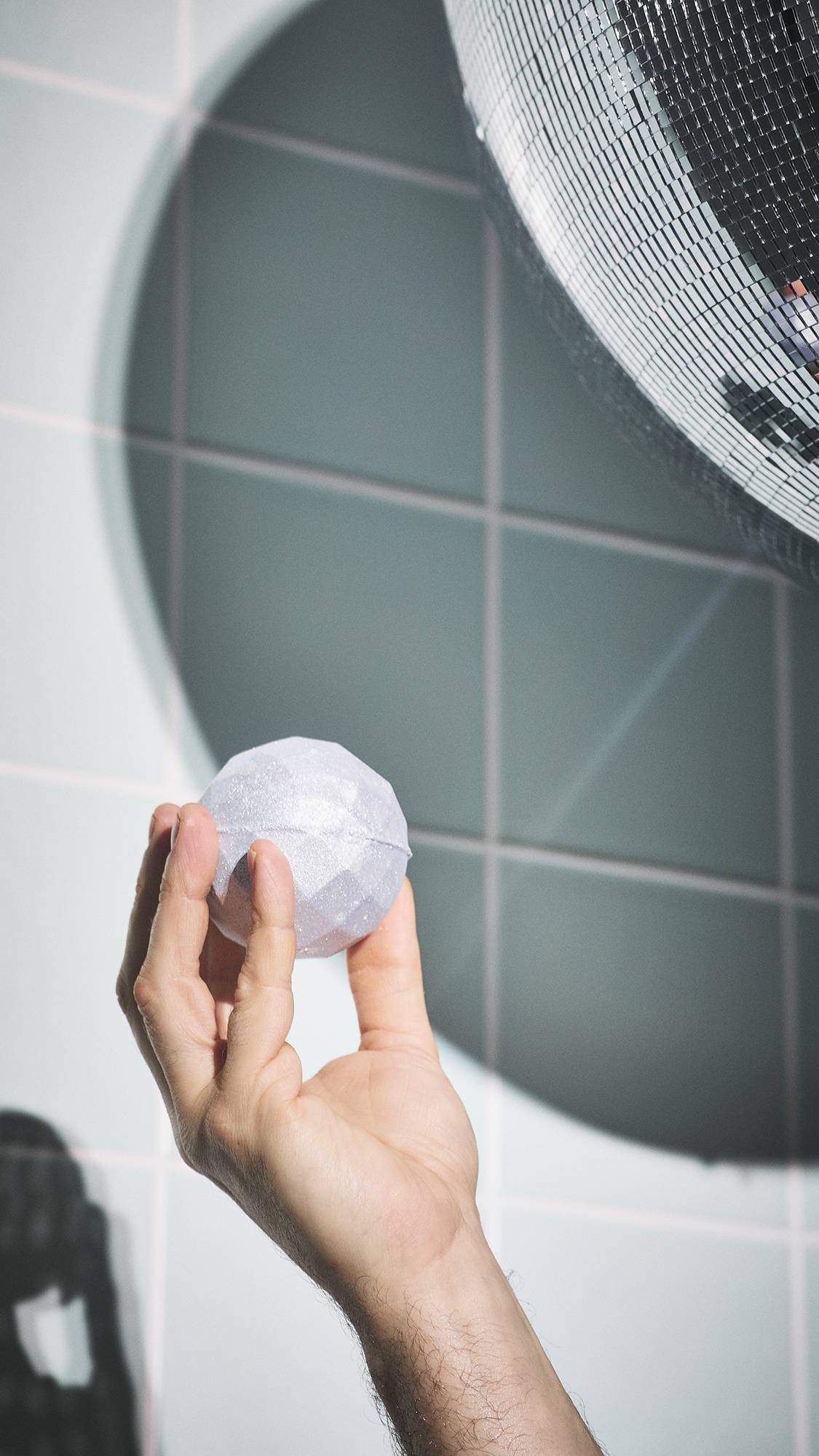 A hand holds a shimmering, "Bathe All The Time" bath bomb with a faceted surface. It is held up towards a large, sparkling disco ball in a bathroom, with a beam of light creating bright reflections and shadows against the pale-tiled wall.