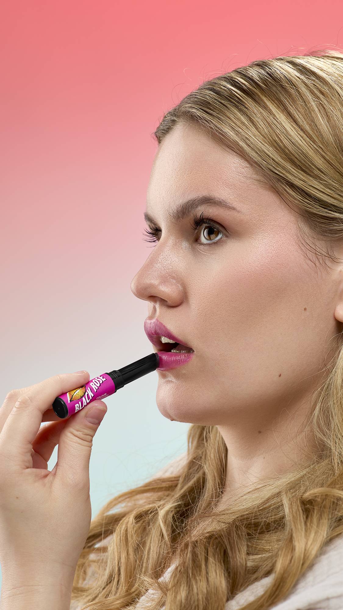 A person with blonde wavy hair applies Black Rose Lip Stain from a pink and black tube. Their lips are tinted a deep berry color. They are shown in profile against a pink-to-blue gradient background.