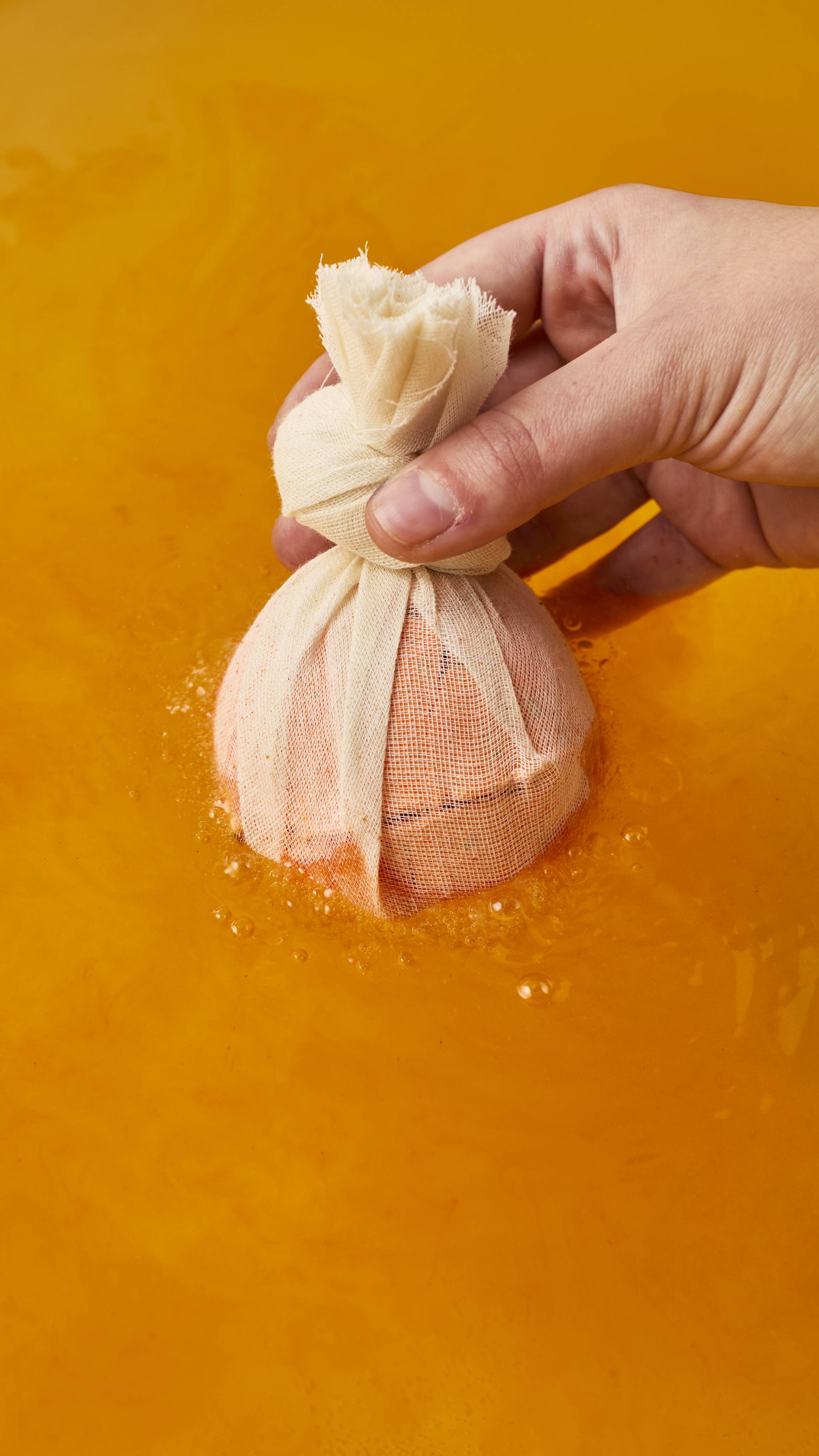 A hand lowers a round bath bomb wrapped in light-coloured cheesecloth into vibrant orange bathwater. Tiny bubbles form around the base of the fabric pouch as it touches the surface.