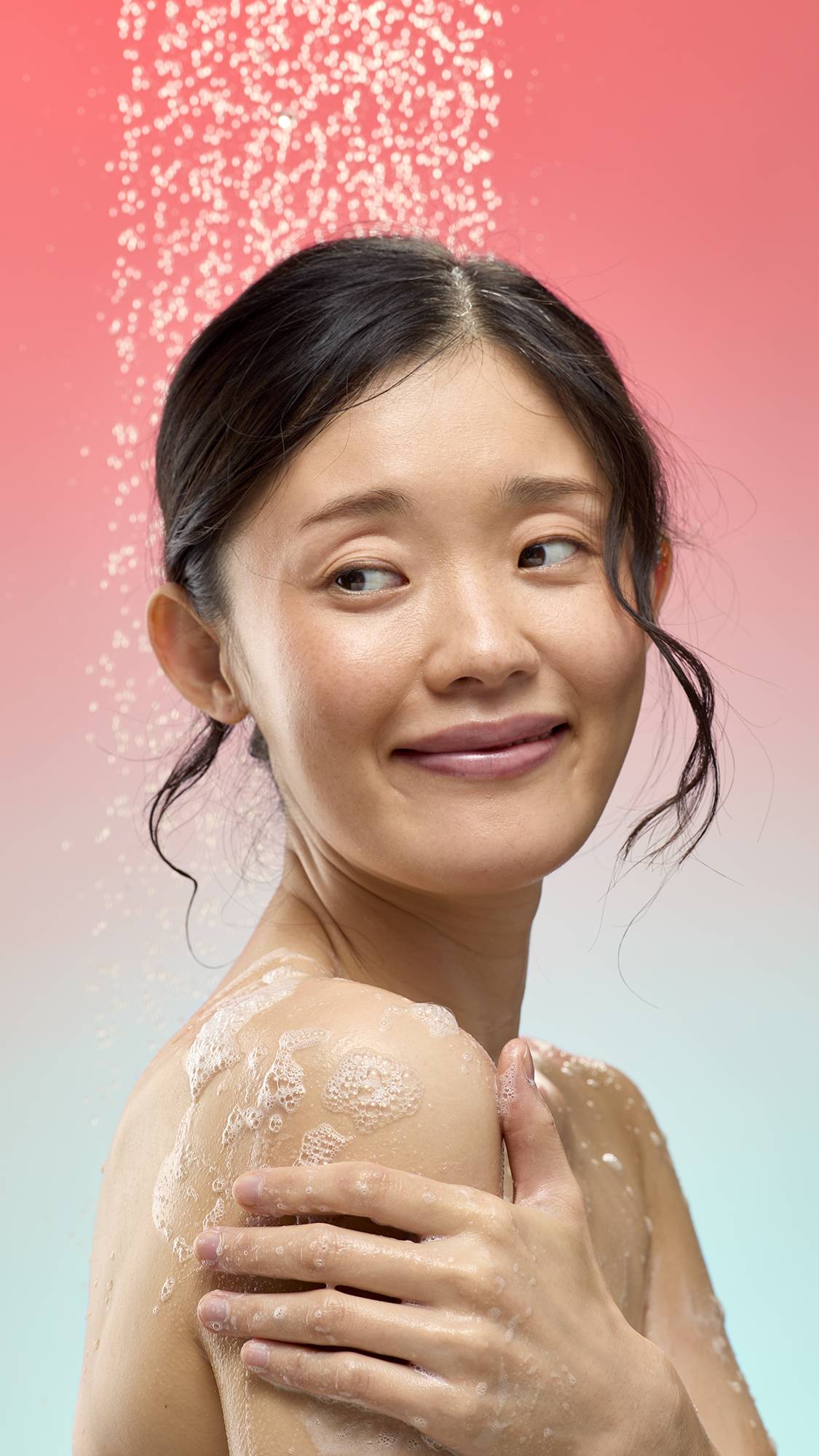 A person smiles while applying foamy white lather from the Bouncing Spider shower gummy to their shoulder. They are standing under a spray of falling water droplets against a soft pink-to-blue gradient background.