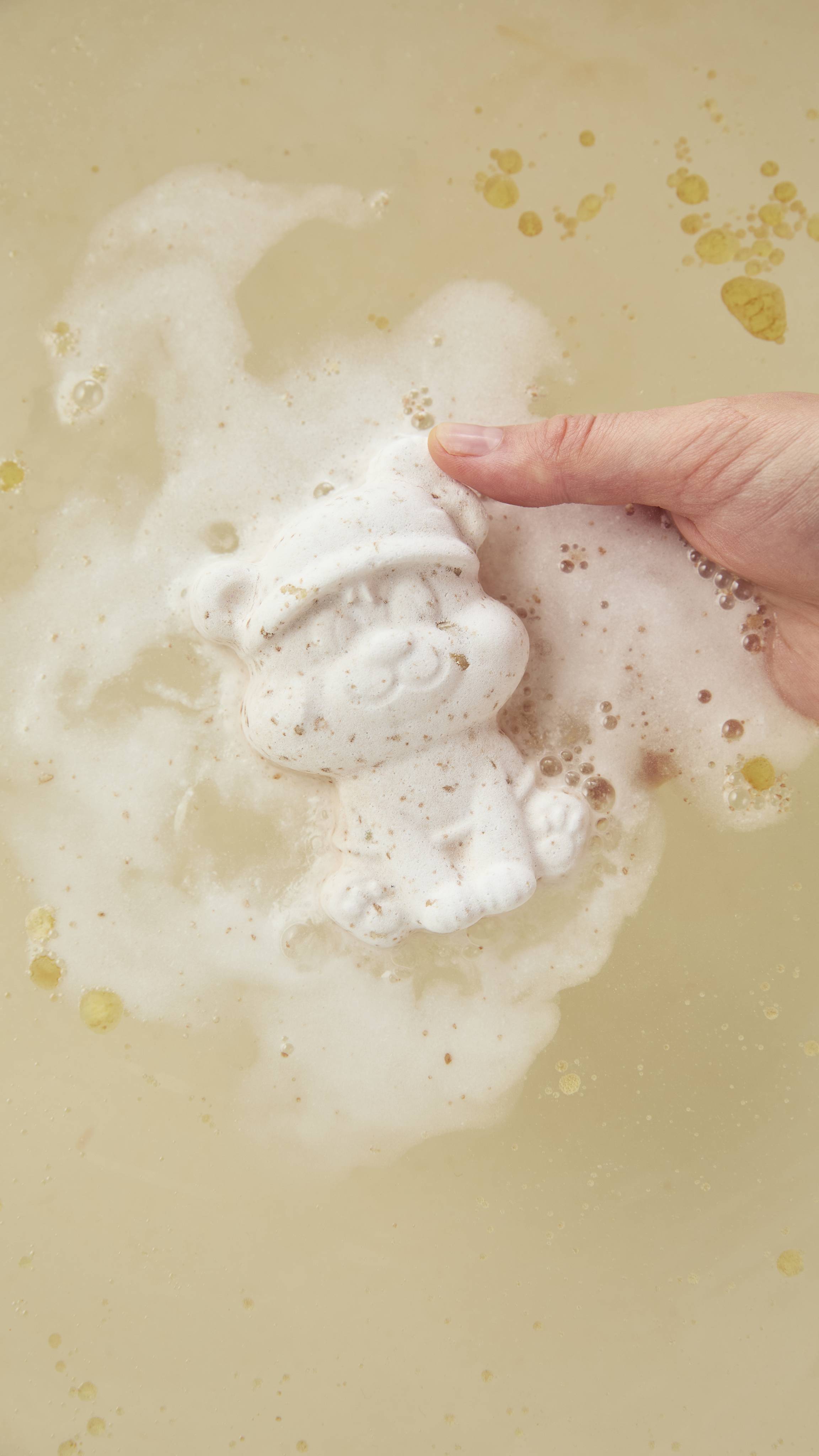 A hand holds a Lush "Butterbear" white polar bear bath bomb wearing a hat and gently places it on light golden bathwater, surrounded by soft white foam