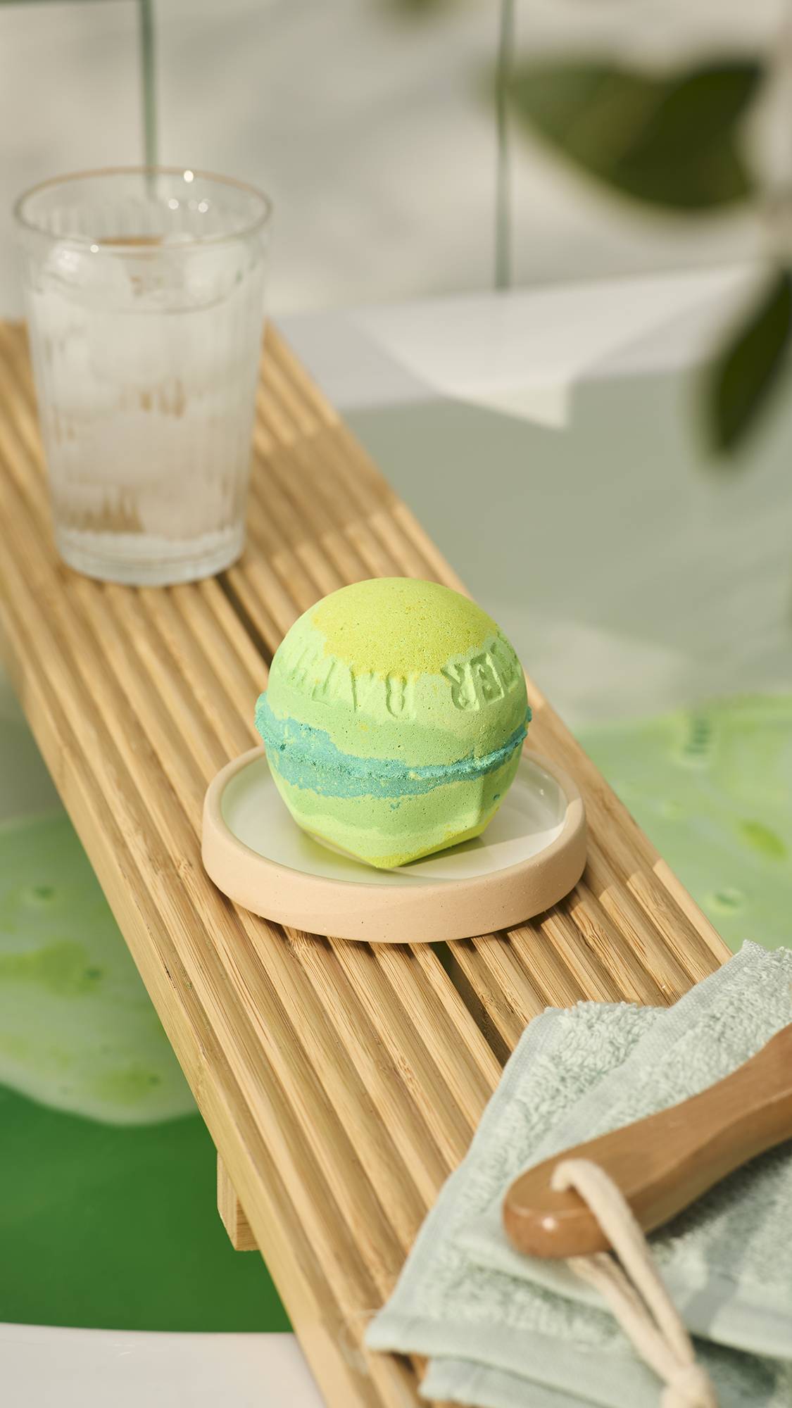 A LUSH "Byegraine" bath bomb with yellow, lime-green, and blue layers sits on a small dish atop a wooden bath caddy, alongside an iced drink and a folded cloth, with green bathwater visible below.