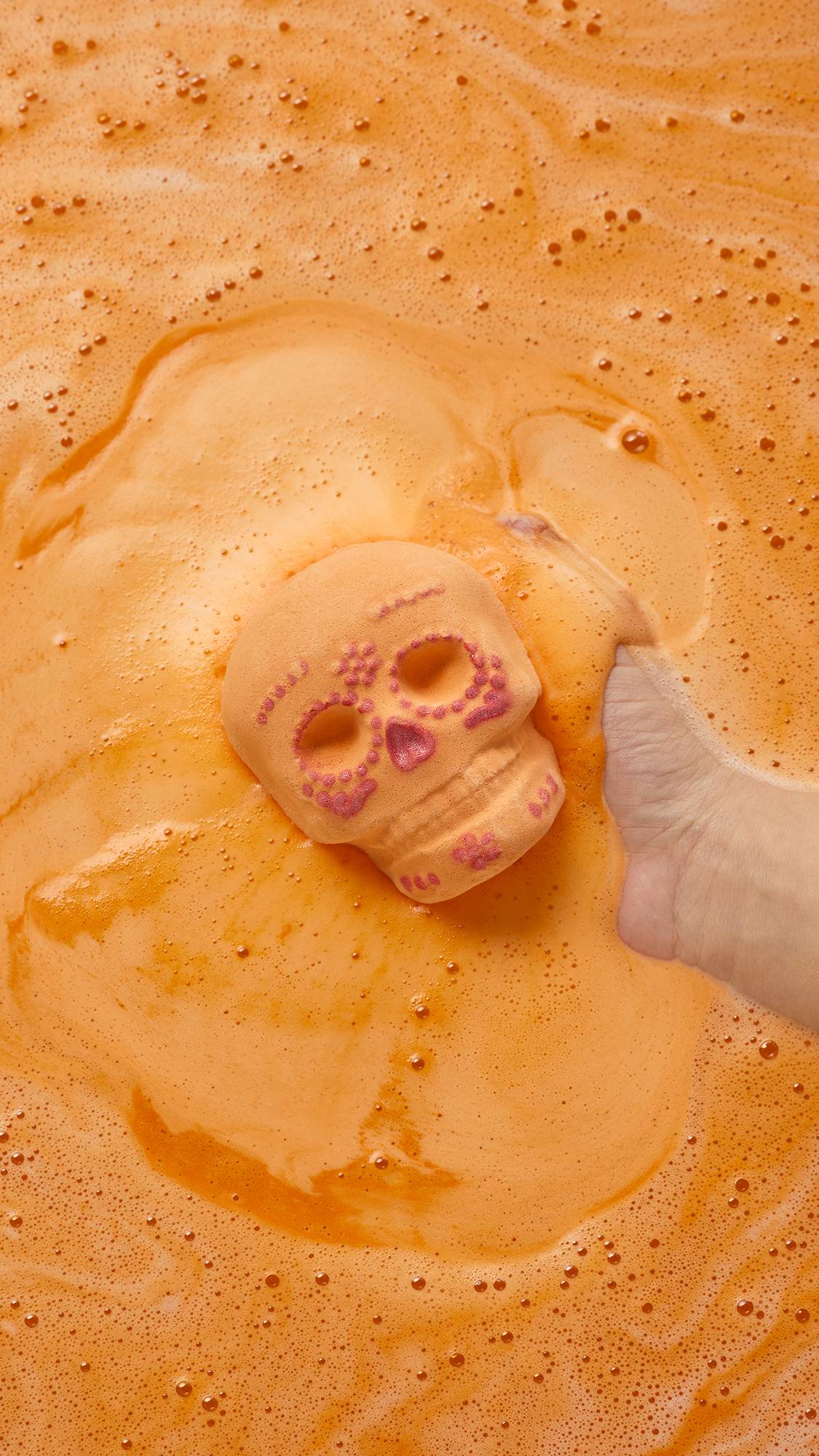 An aerial view of the Lush Calaverita sugar skull-inspired bath bomb as a hand can be seen slowly lowering the product into the water, surrounded by thick, velvety, orange foam.