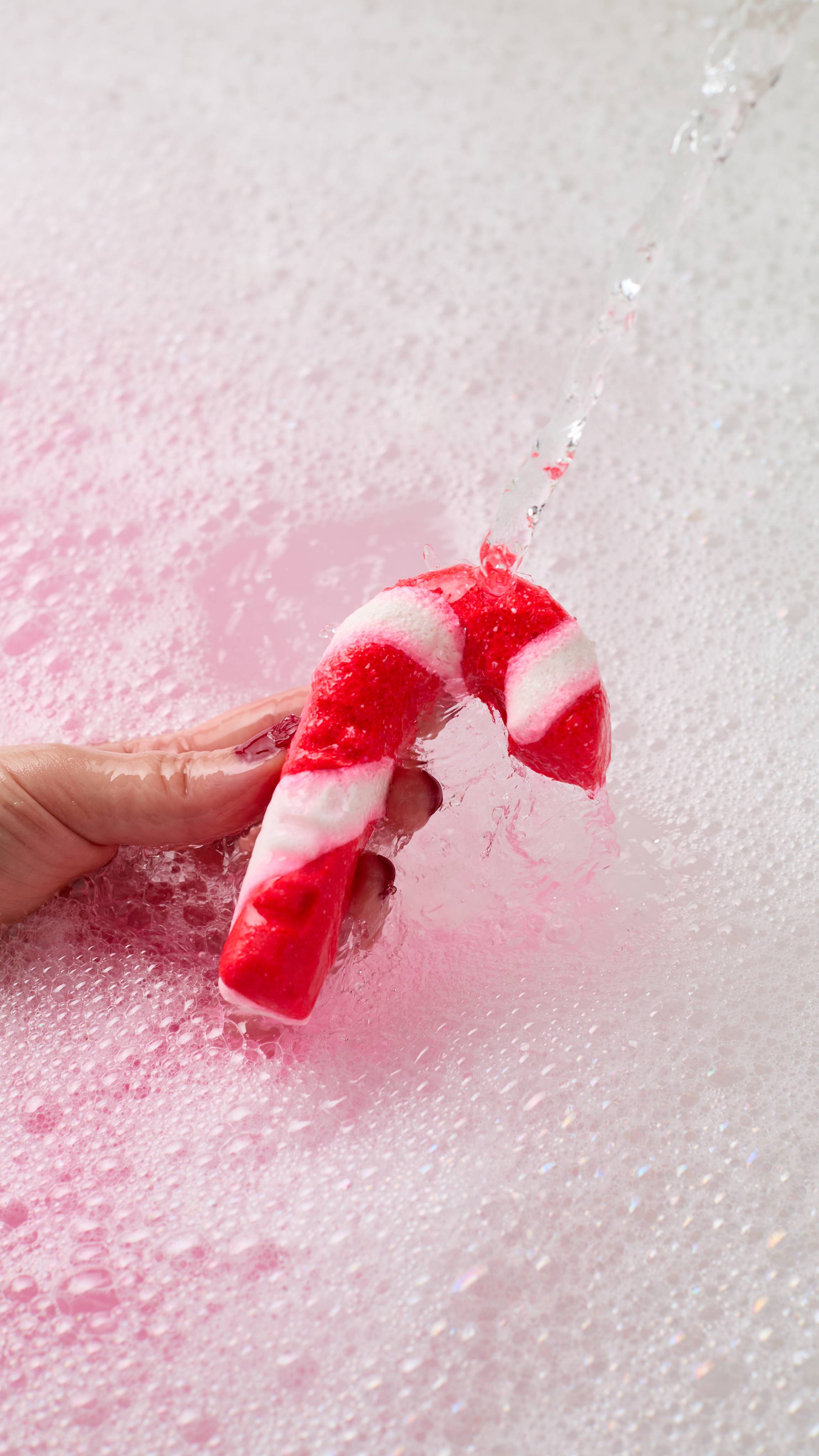 A hand holds a red and white striped candy cane bubble bar under running tap water, creating a sea of white bubbles and soft pink foam in the bathwater.