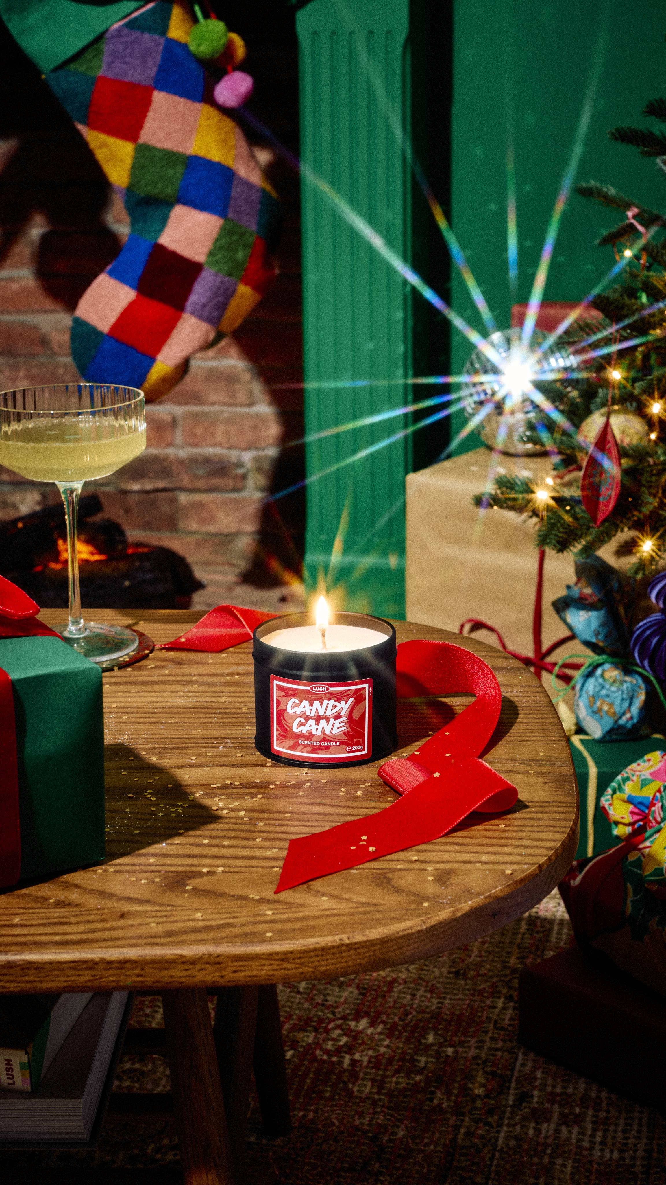 A lit tin of Lush Candy Cane scented candle burns on a wooden table beside red ribbon, a cocktail glass, and a wrapped gift, set in a festive room with a Christmas tree and colourful stocking.