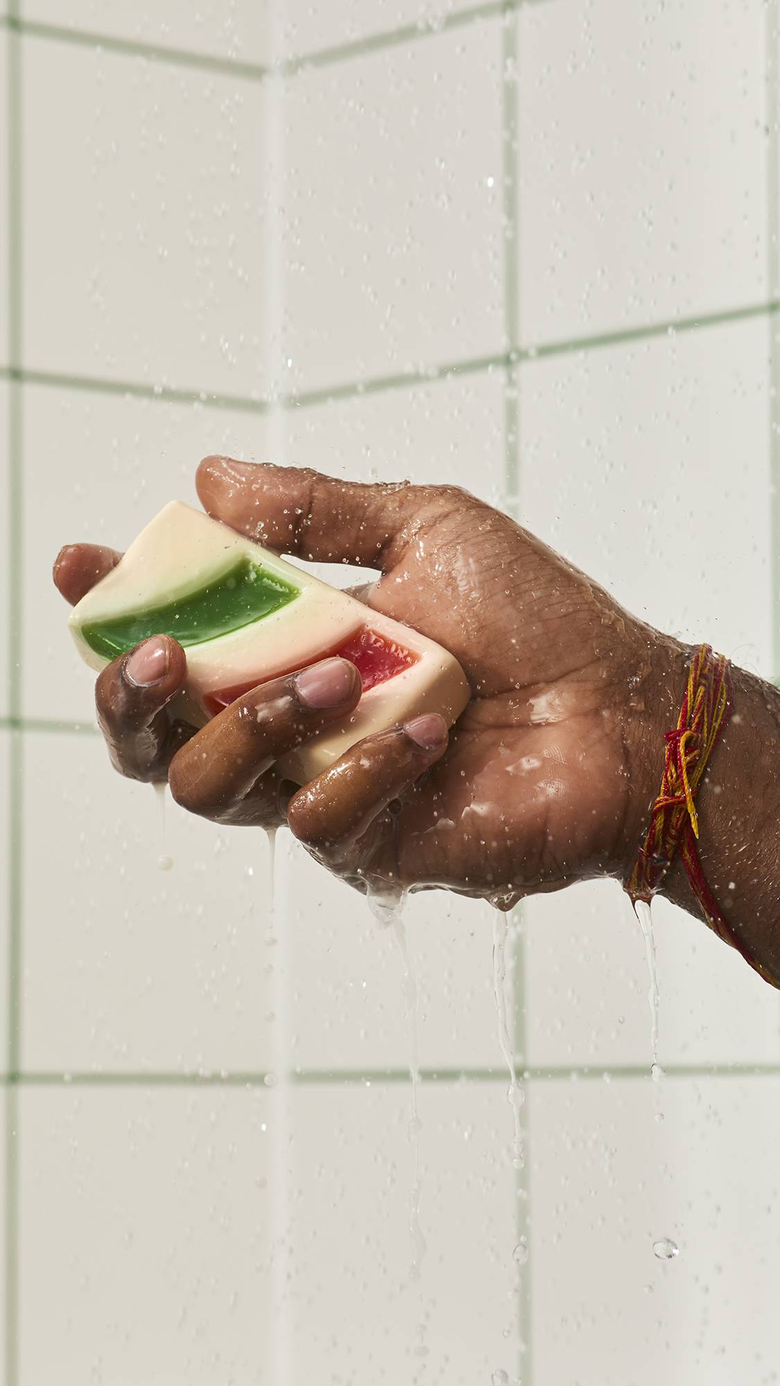A hand holds a bar of cream-coloured "Candy Cane" soap with green and red striped accents under streaming shower water, set against a white tiled wall background.