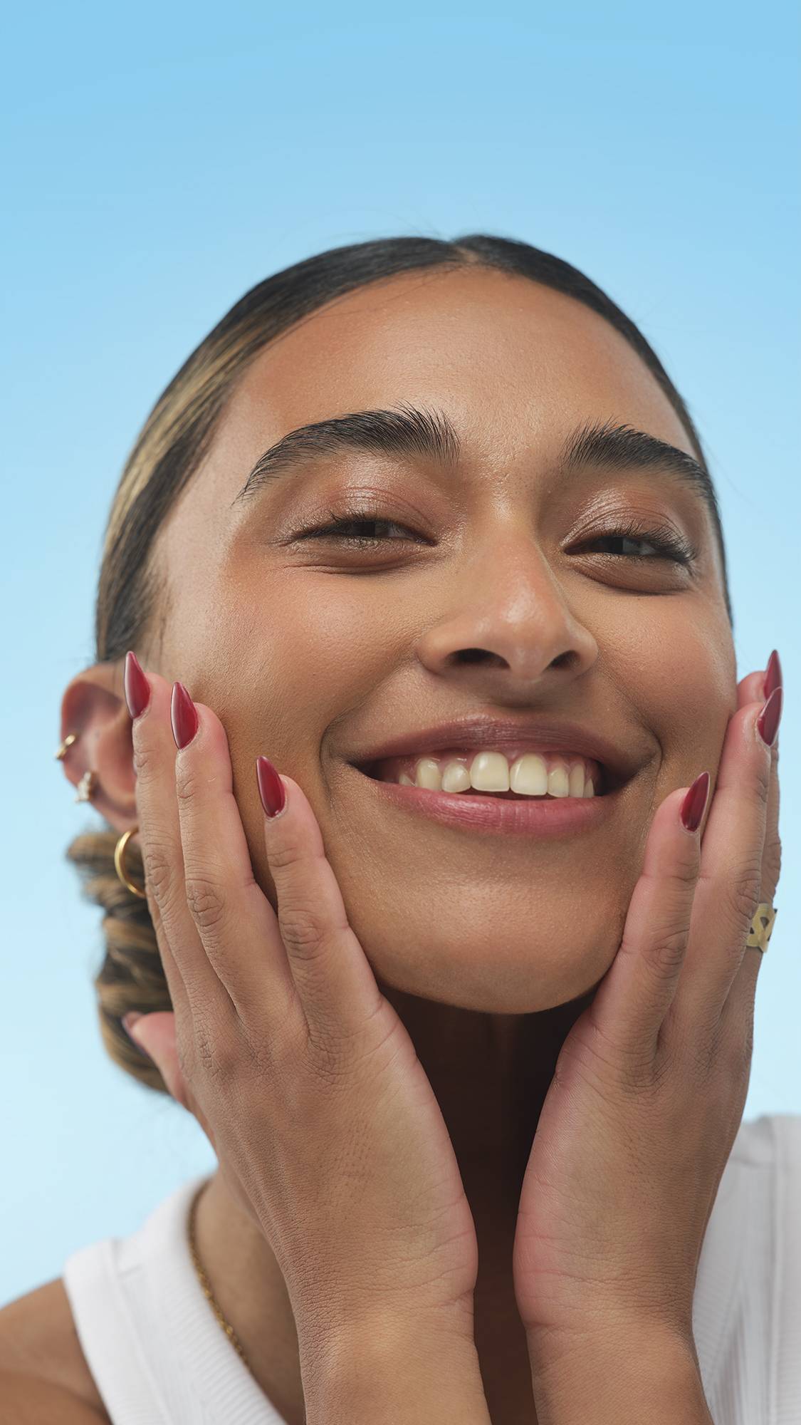 A close-up portrait of a broadly smiling person with warm-toned skin. They cup their cheeks with hands, showing dark red nails and a bronzed, radiant glow from the Charisma Skin tint, against a bright blue background.