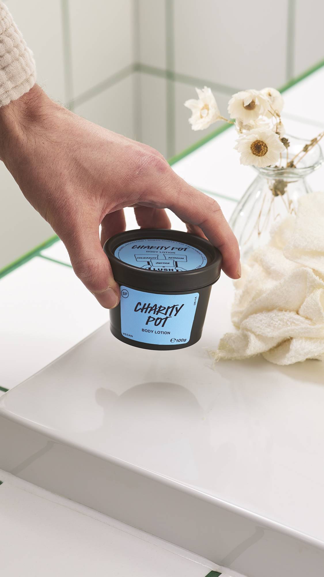A hand places a small black tub of Lush Charity Pot body lotion on a glossy white surface, beside a small vase of dried white flowers and a folded flannel against a tiled bathroom wall.