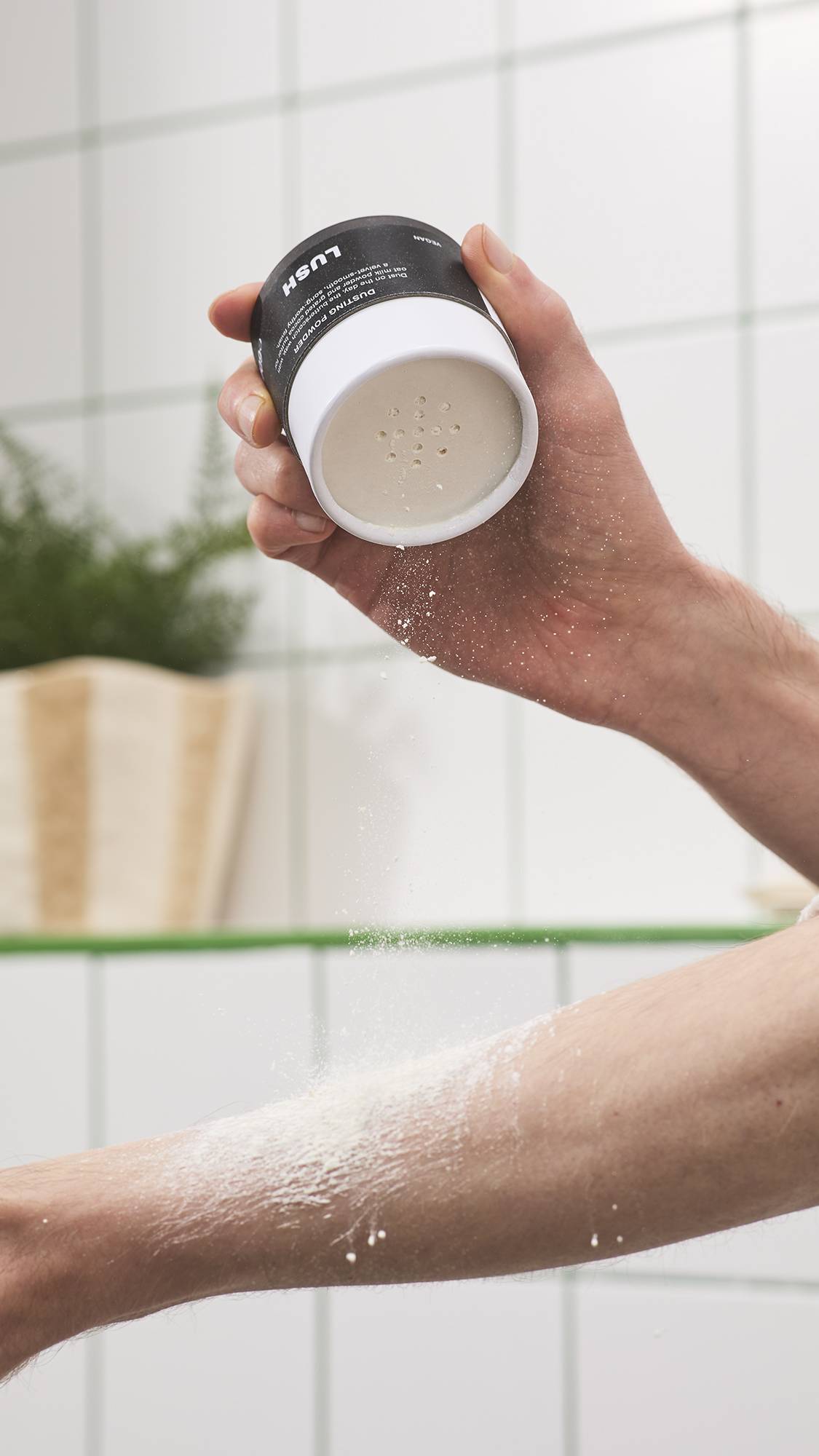 A hand shakes fine white Lush "Chelsea Morning" dusting powder from a black tub with shaker holes onto an arm, leaving a light layer of powder across the skin against a white tiled bathroom background.