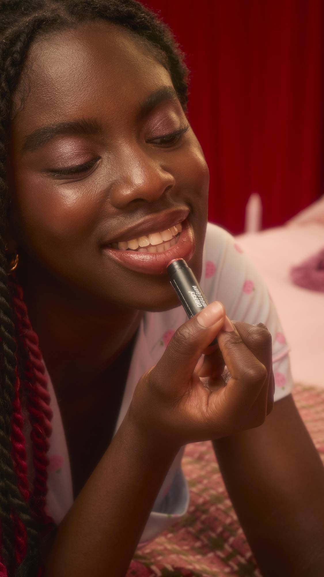 A person wearing strawberry-patterned pyjamas smiles while applying a reddish-pink Lush "Cherry Bubblegum" solid lip oil to their bottom lip. They are reclining on a patterned bedspread in a warm, dimly lit room.